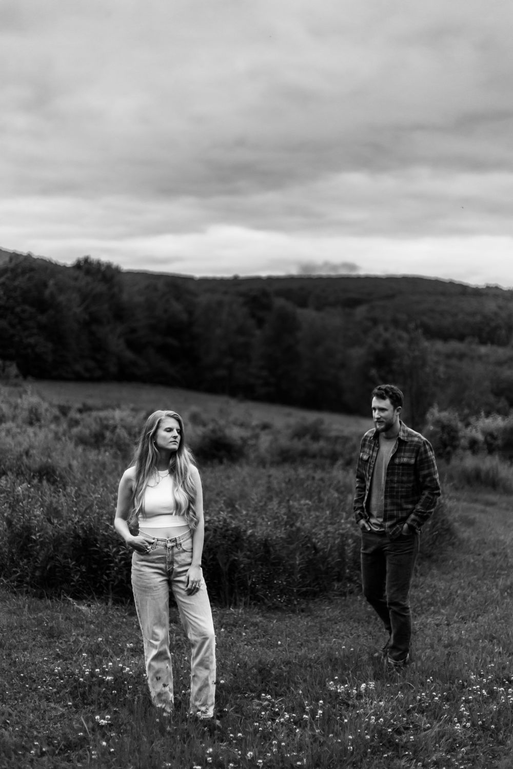 Black and white photo of a couple standing apart in a lush meadow with rolling hills and cloudy sky.