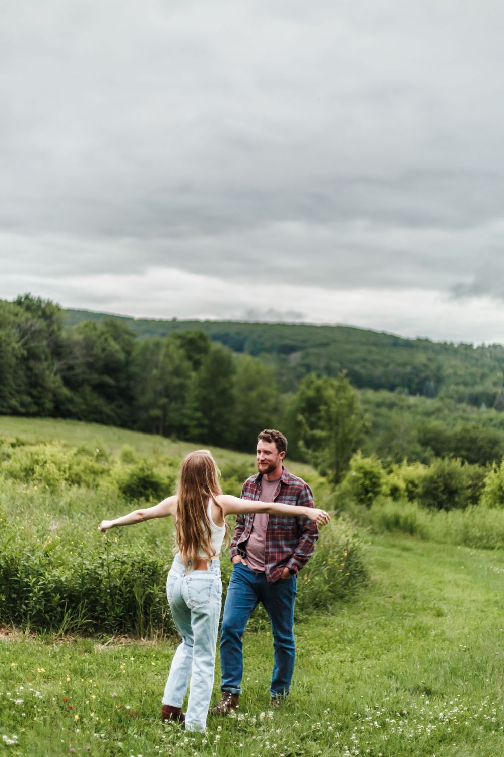 Couple laughing together in a green meadow with rolling hills and trees under a cloudy sky.