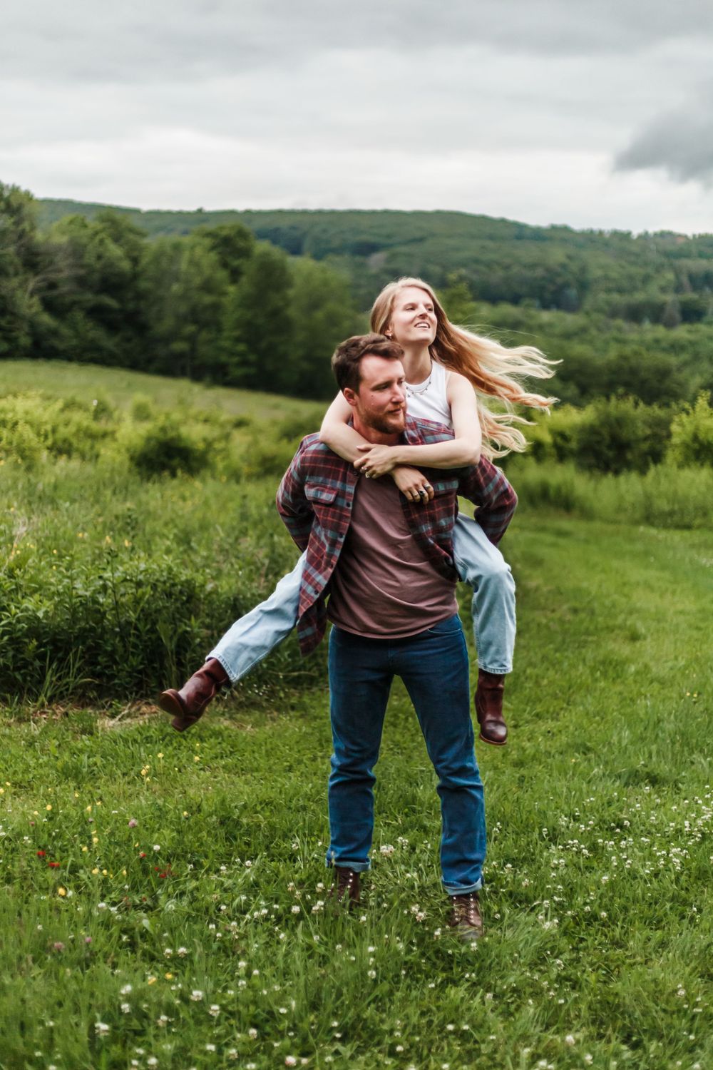 Man giving woman a piggyback ride in a lush green meadow with rolling hills in the background.