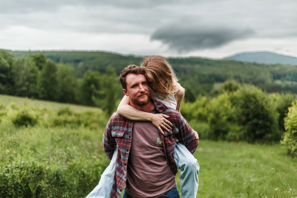 Couple sharing a piggyback ride in a lush green field with mountains in the background on a cloudy day.