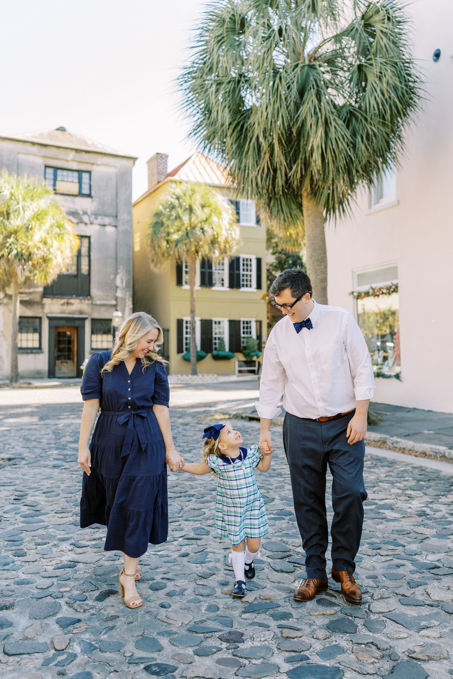 Family of three walking on cobblestone street in Charleston, SC, with palm trees and colorful historic buildings.