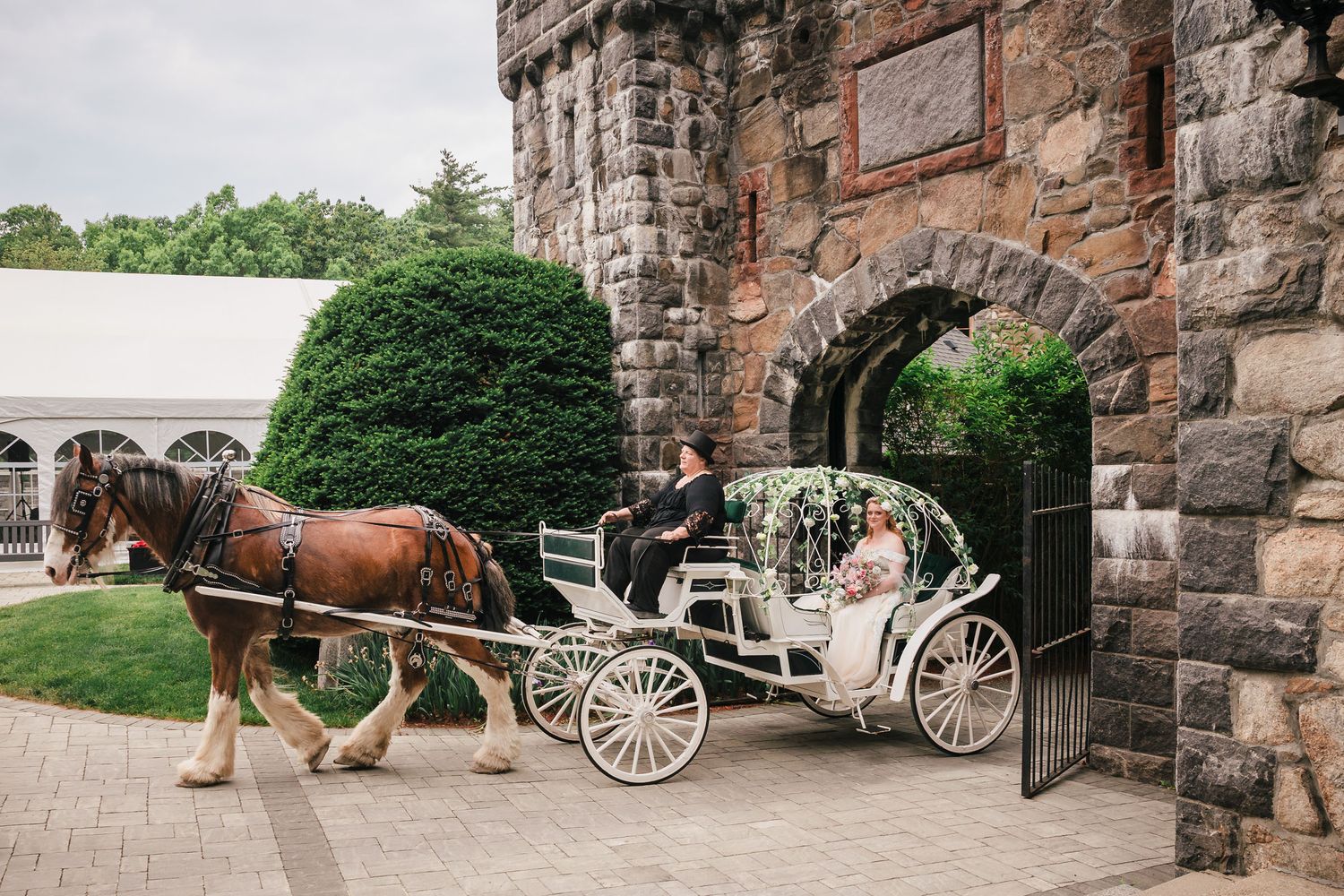 A draft horse pulling the bride's rose-covered carriage past Searles Castle's stone archway.