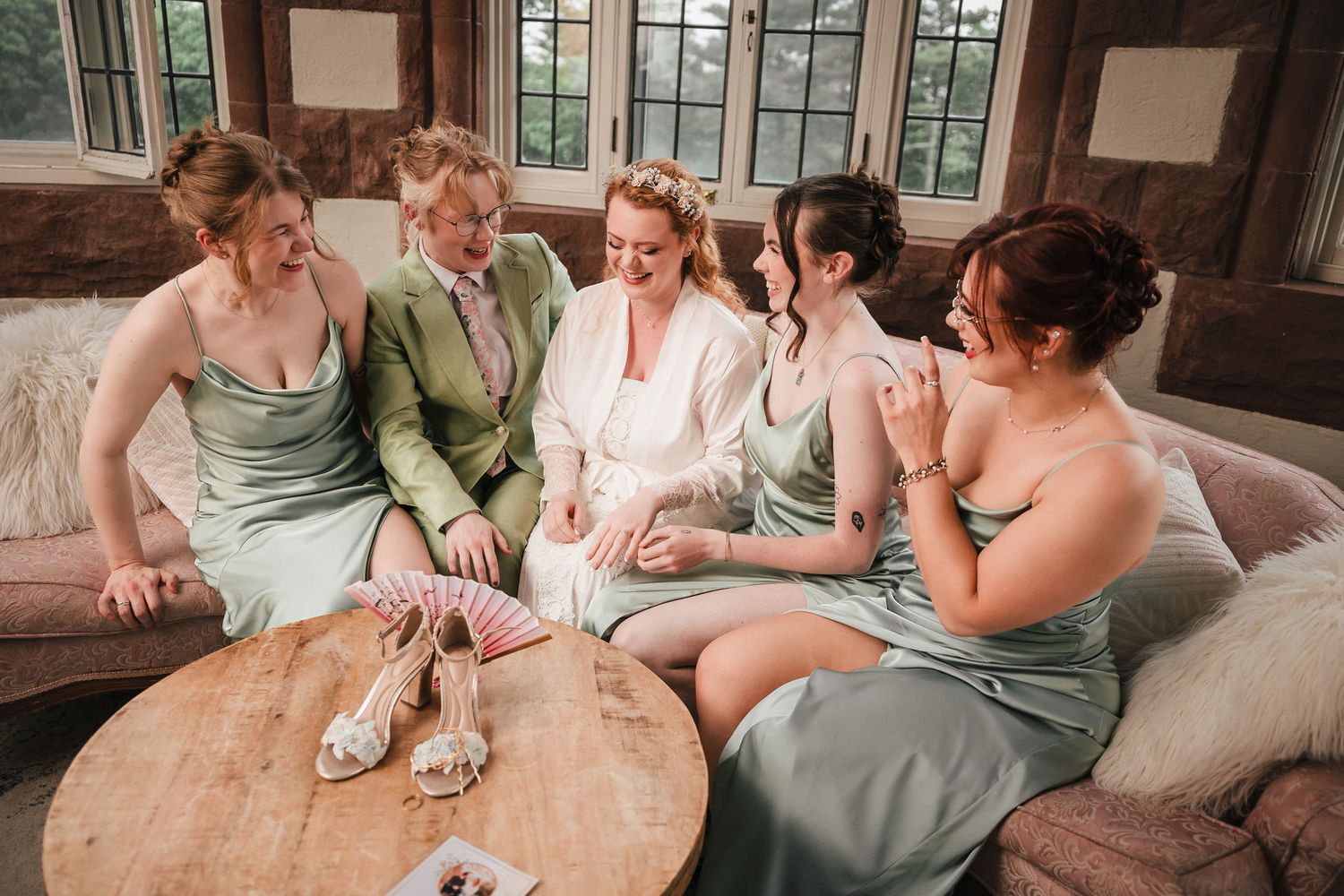 Bride in white robe laughing with bridesmaids in sage green dresses on a couch at a Searless Castle.