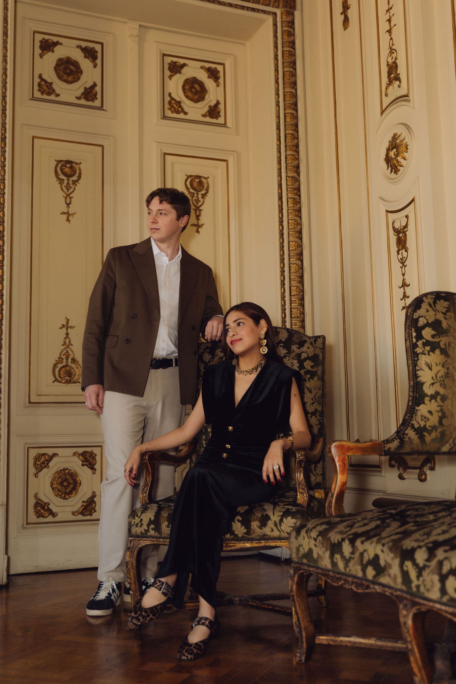 Elegant couple posing in ornate French-style room, man standing beside woman seated in antique chair.