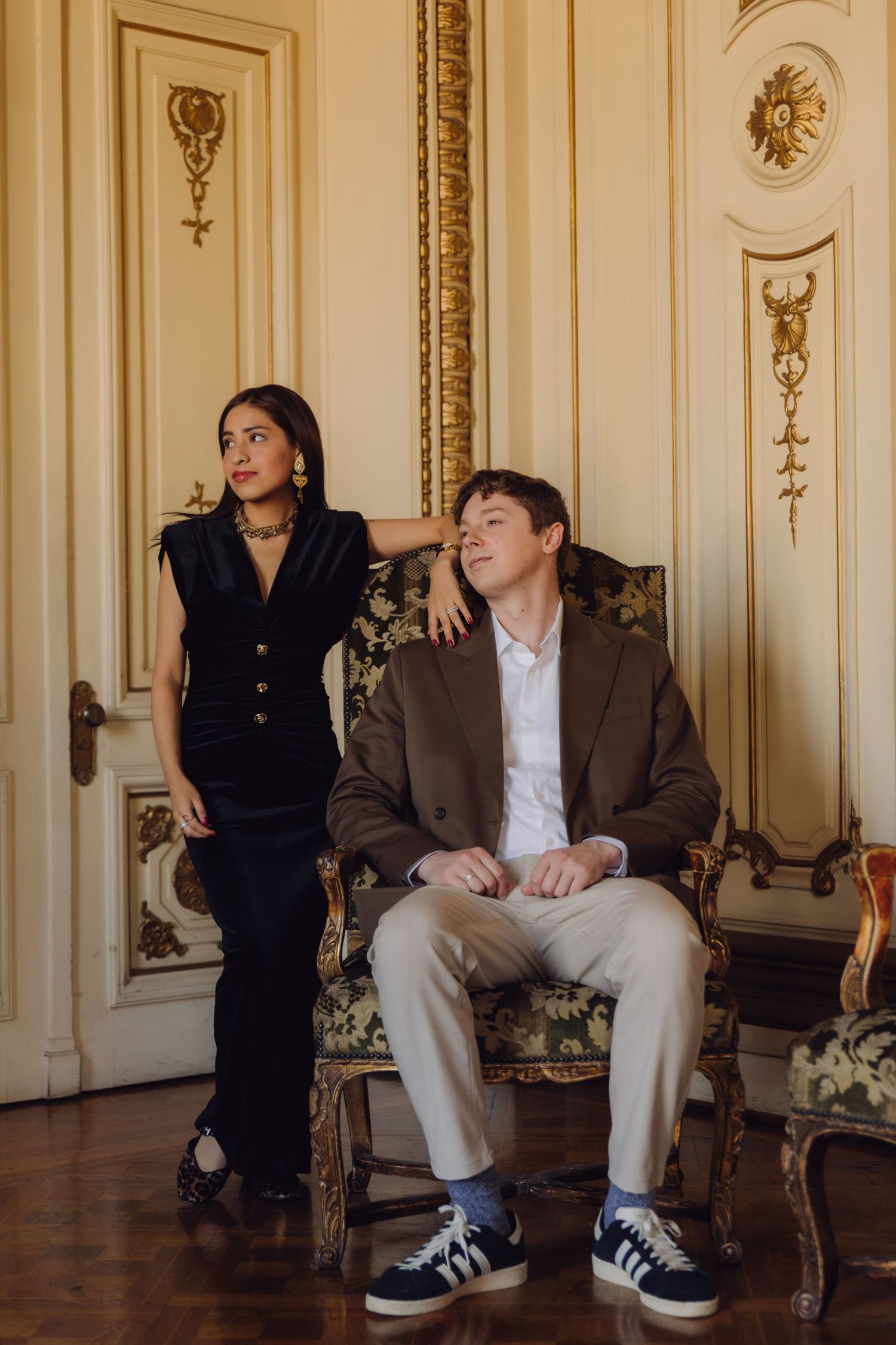 Stylish couple in luxurious baroque interior, woman in black outfit standing beside seated man in brown blazer.