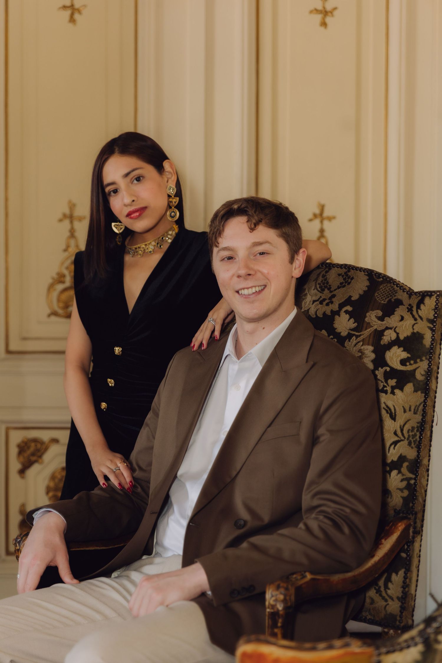 Smiling couple posing in elegant room, woman in black dress standing beside man seated in ornate chair.