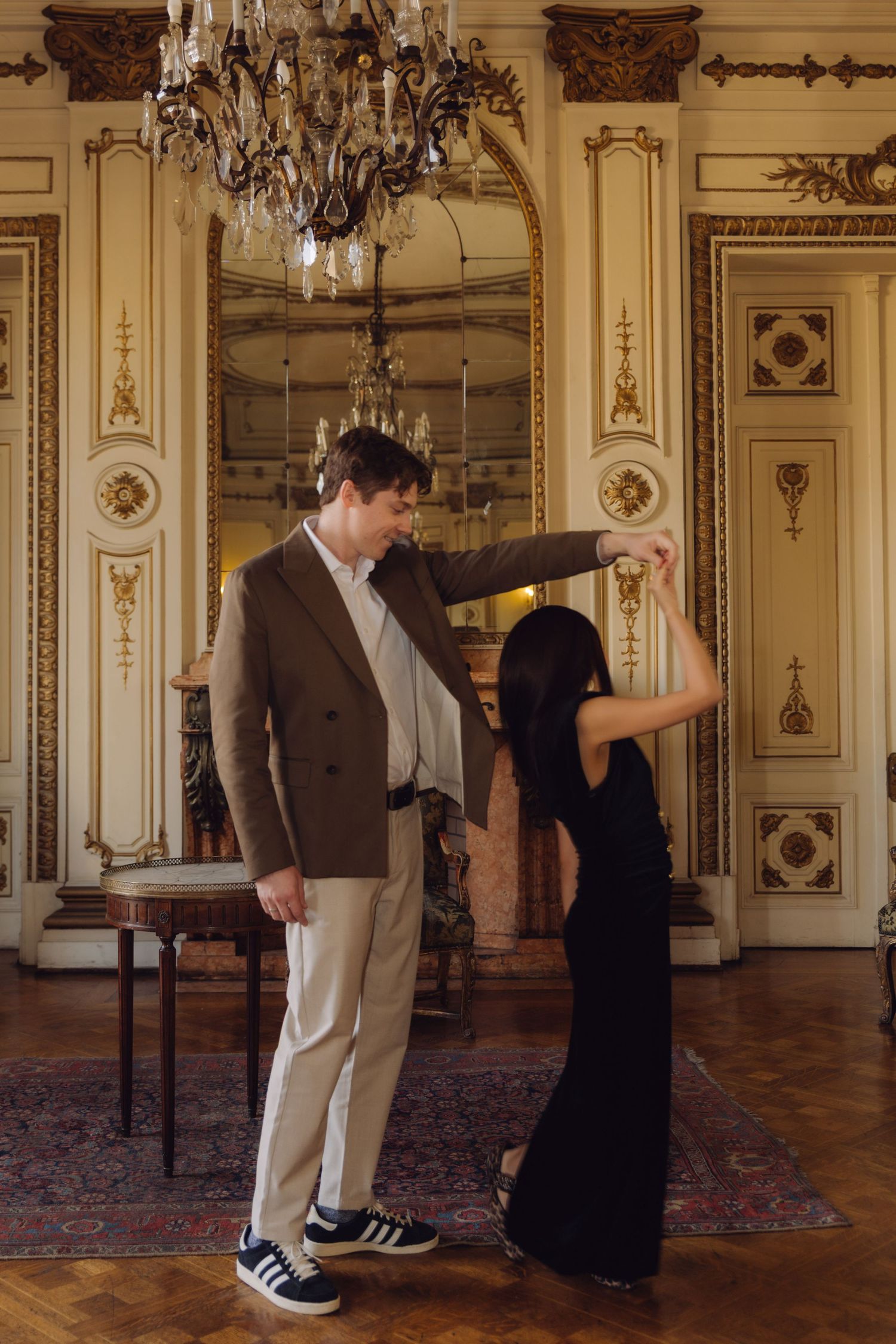 Couple dancing gracefully in opulent chandelier-lit ballroom, man in brown blazer spinning woman in black dress.