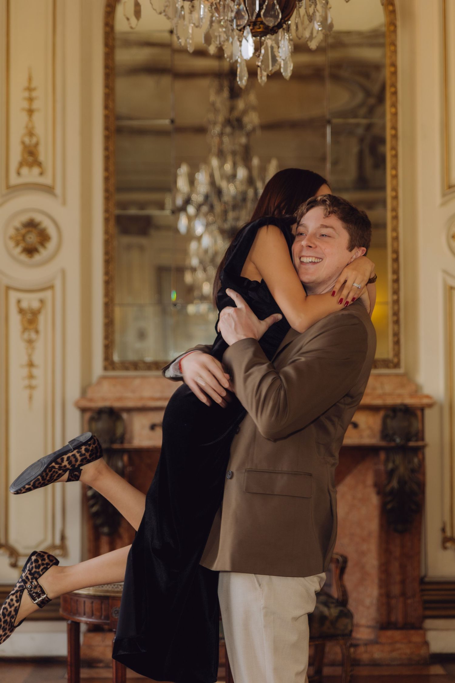 Joyful man in brown blazer lifting smiling woman in black dress in an elegant chandelier-lit room.