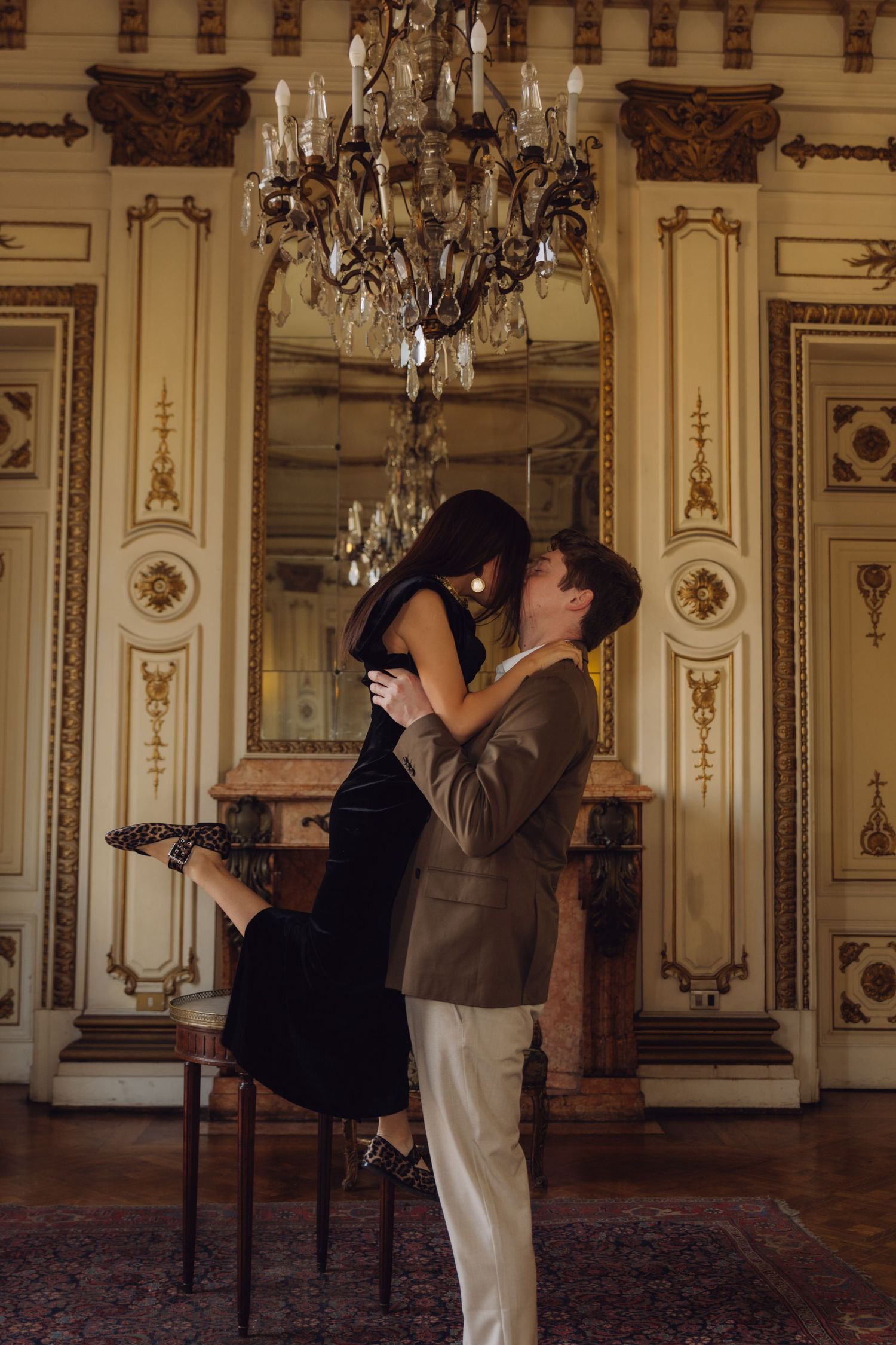 Romantic couple sharing a kiss as man lifts woman in black dress beneath a crystal chandelier.