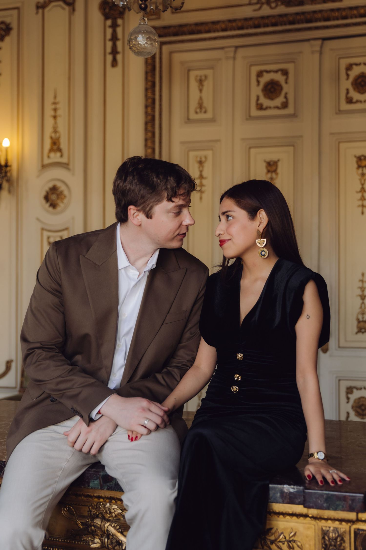 Elegant couple in black dress and brown blazer sitting together in ornate room with gold decor.