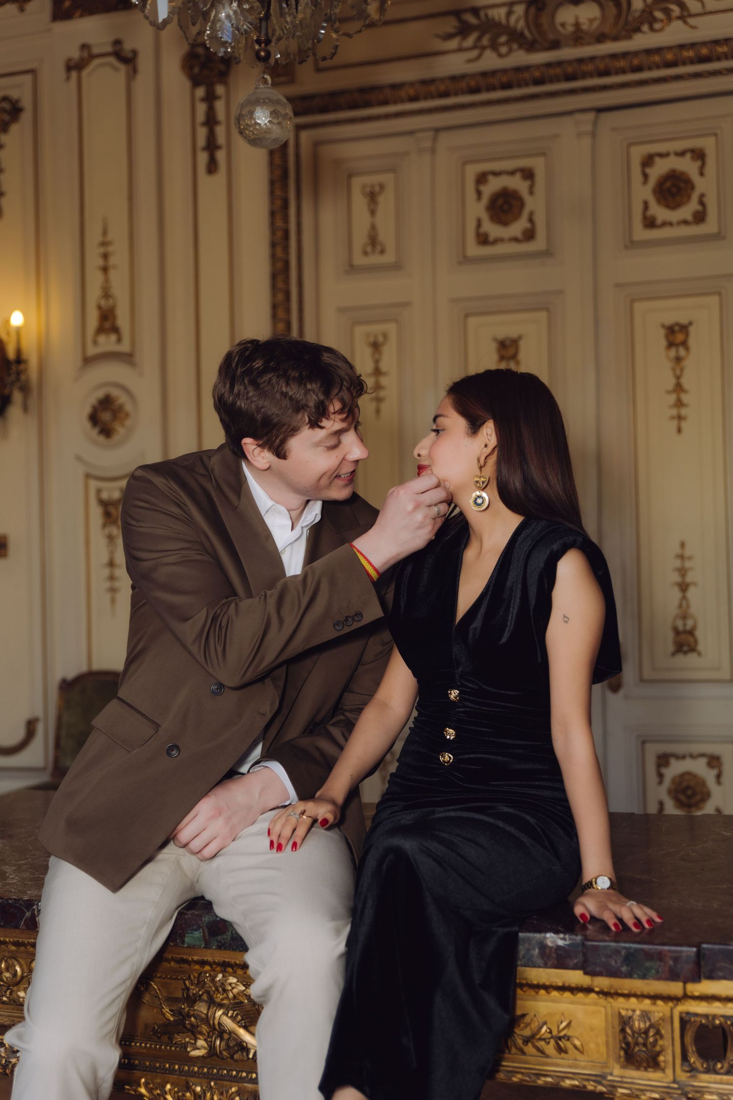 Man in brown blazer gently touching womans face in elegant gold-trimmed classical interior.