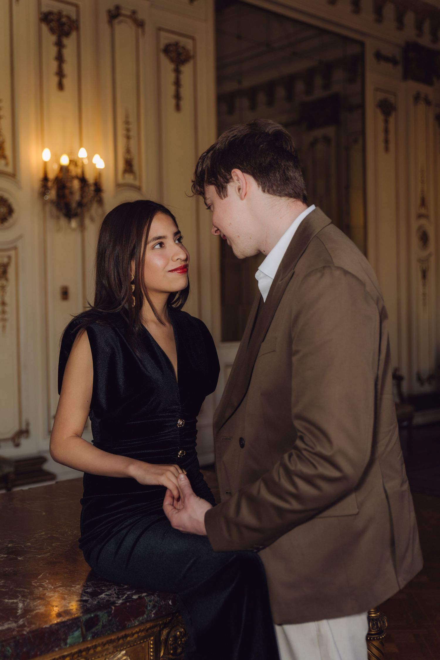 Stylish couple in black dress and brown blazer sharing an intimate gaze in opulent interior.