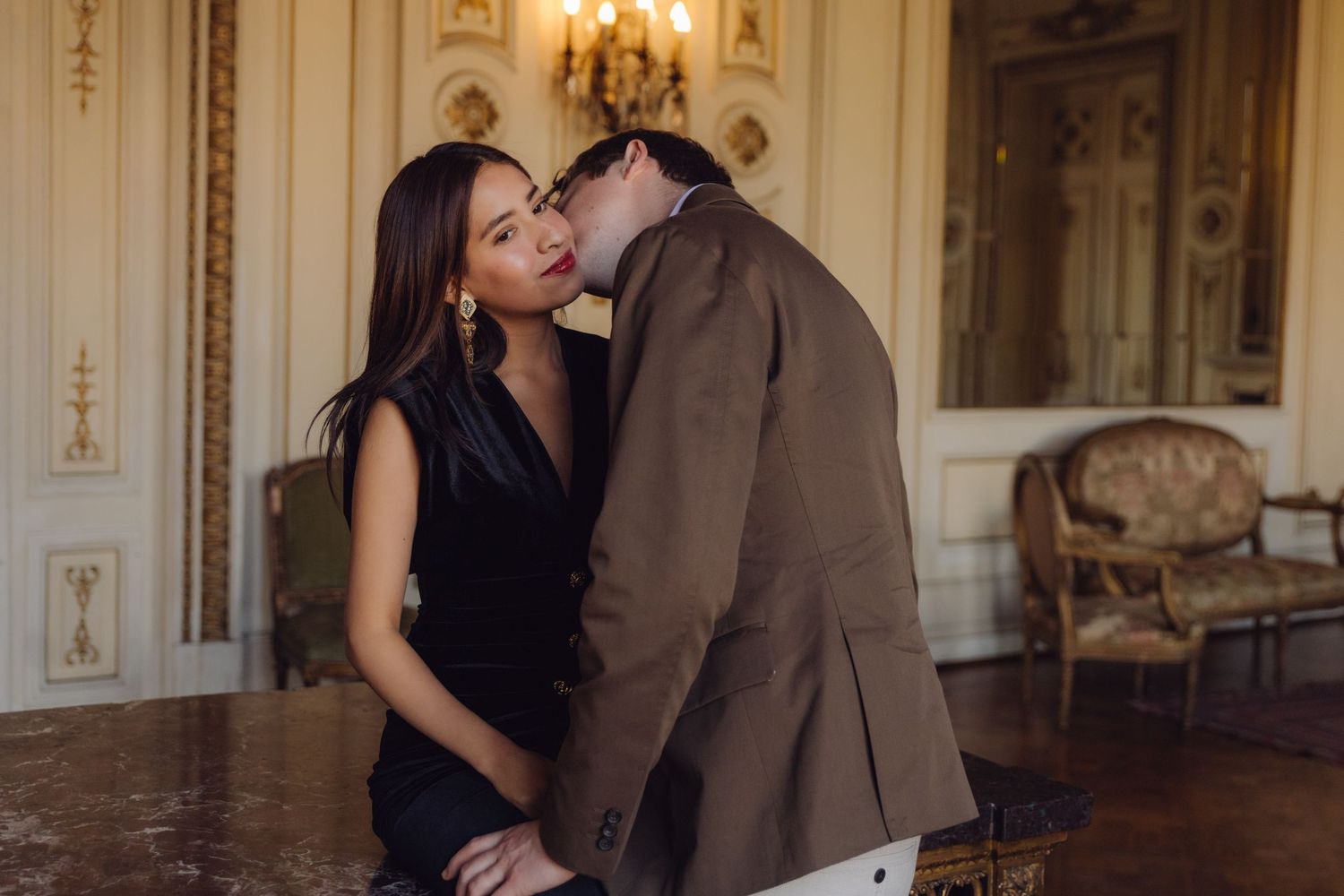 Couple sharing a romantic kiss in a grand classical French-style room with ornate white walls.