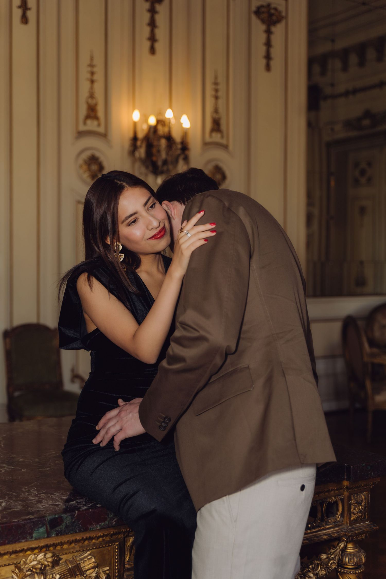 Woman in black velvet dress embracing man in brown blazer in a luxurious baroque-style room.