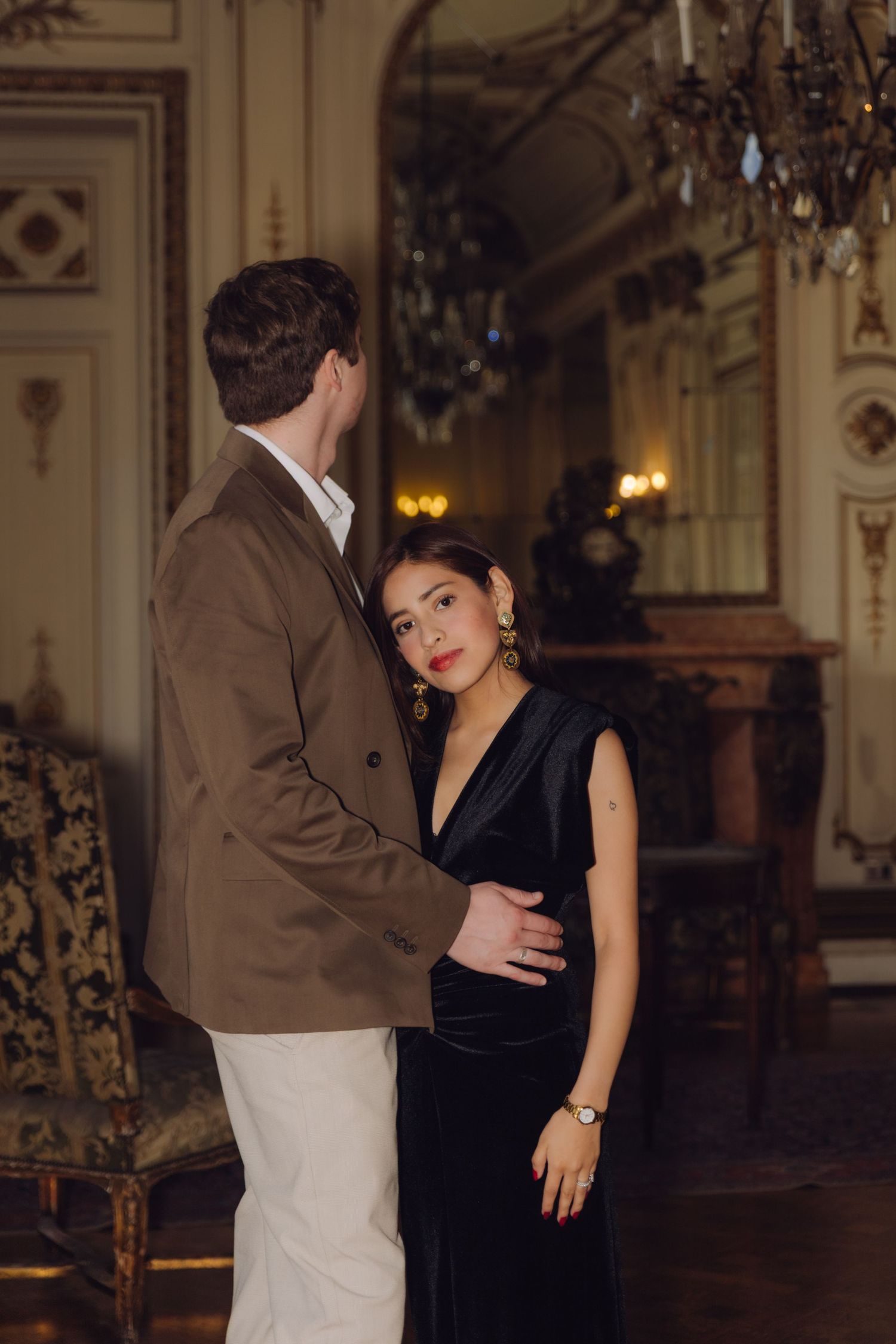 Couple posing in a dimly lit ornate room, woman in black velvet dress with gold earrings.