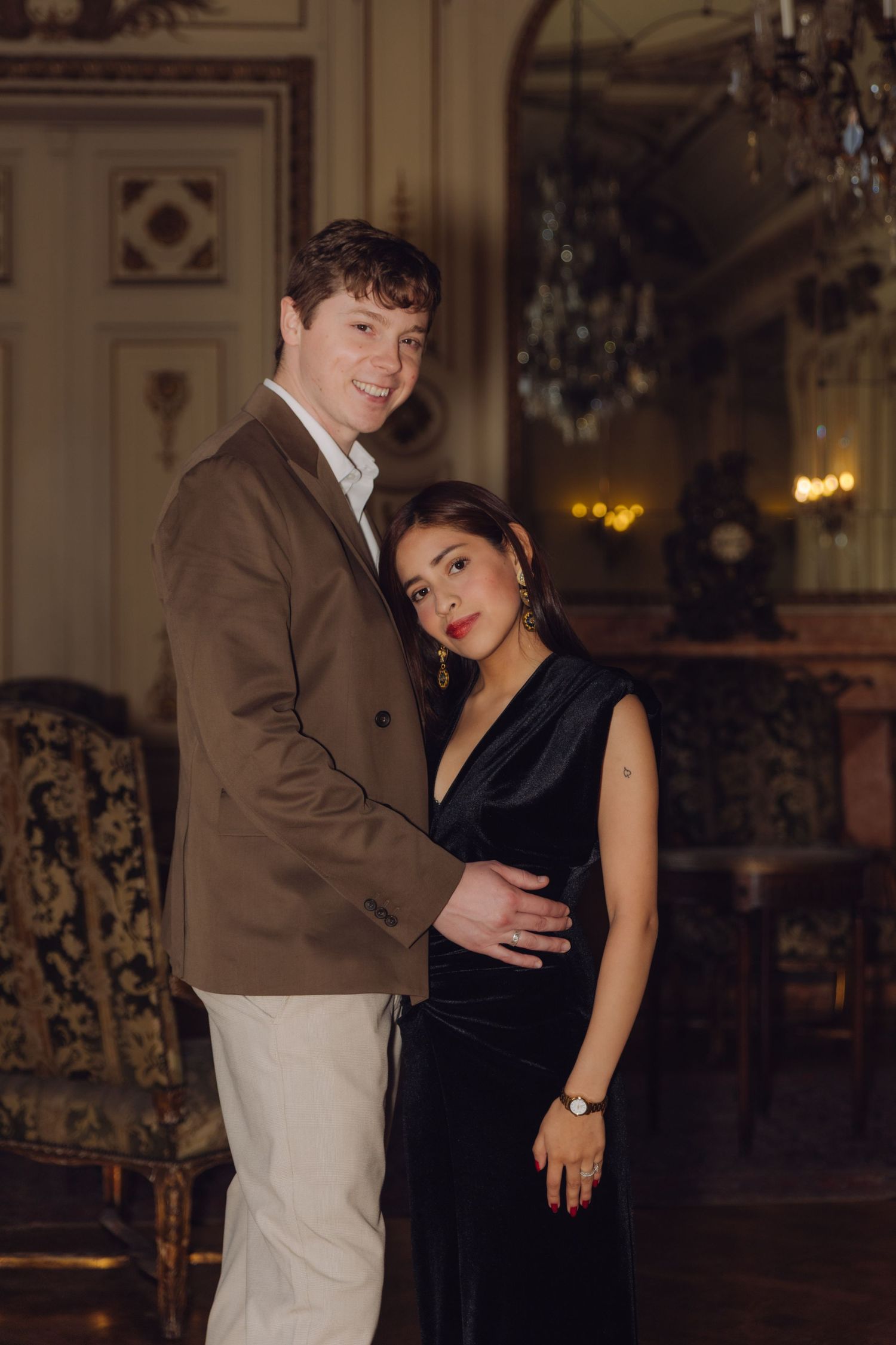 Smiling couple embracing in a grand classical interior with chandelier and vintage furnishings.