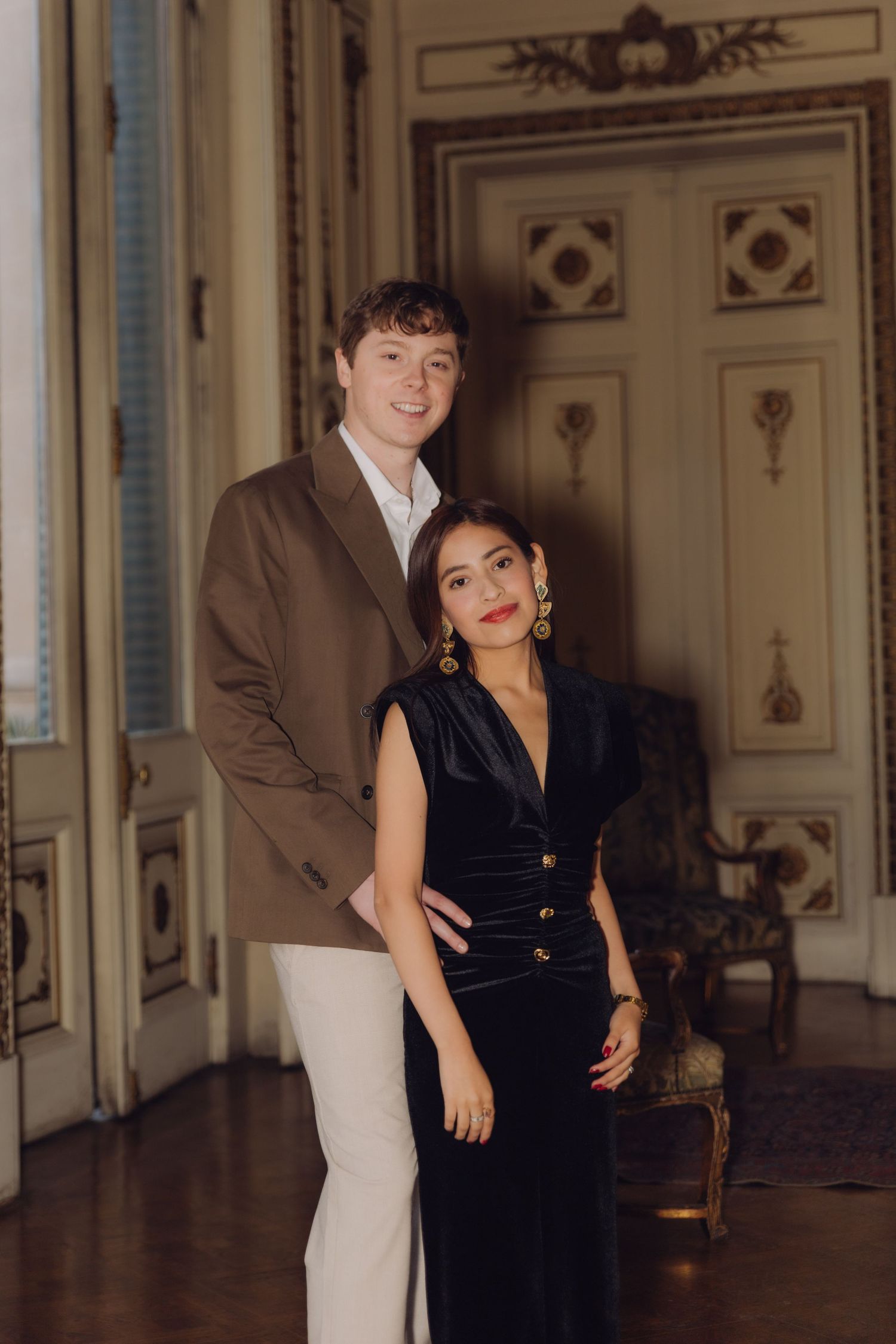 Elegant couple posing in ornate room, man in brown blazer and woman in black velvet dress.