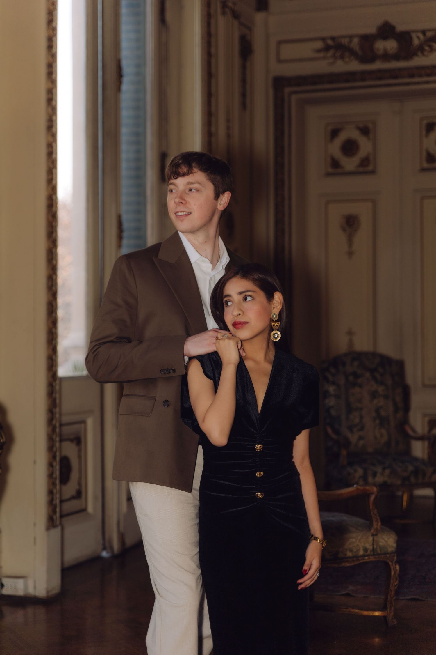Stylish couple standing by window in classic interior, woman in black velvet dress with gold earrings.