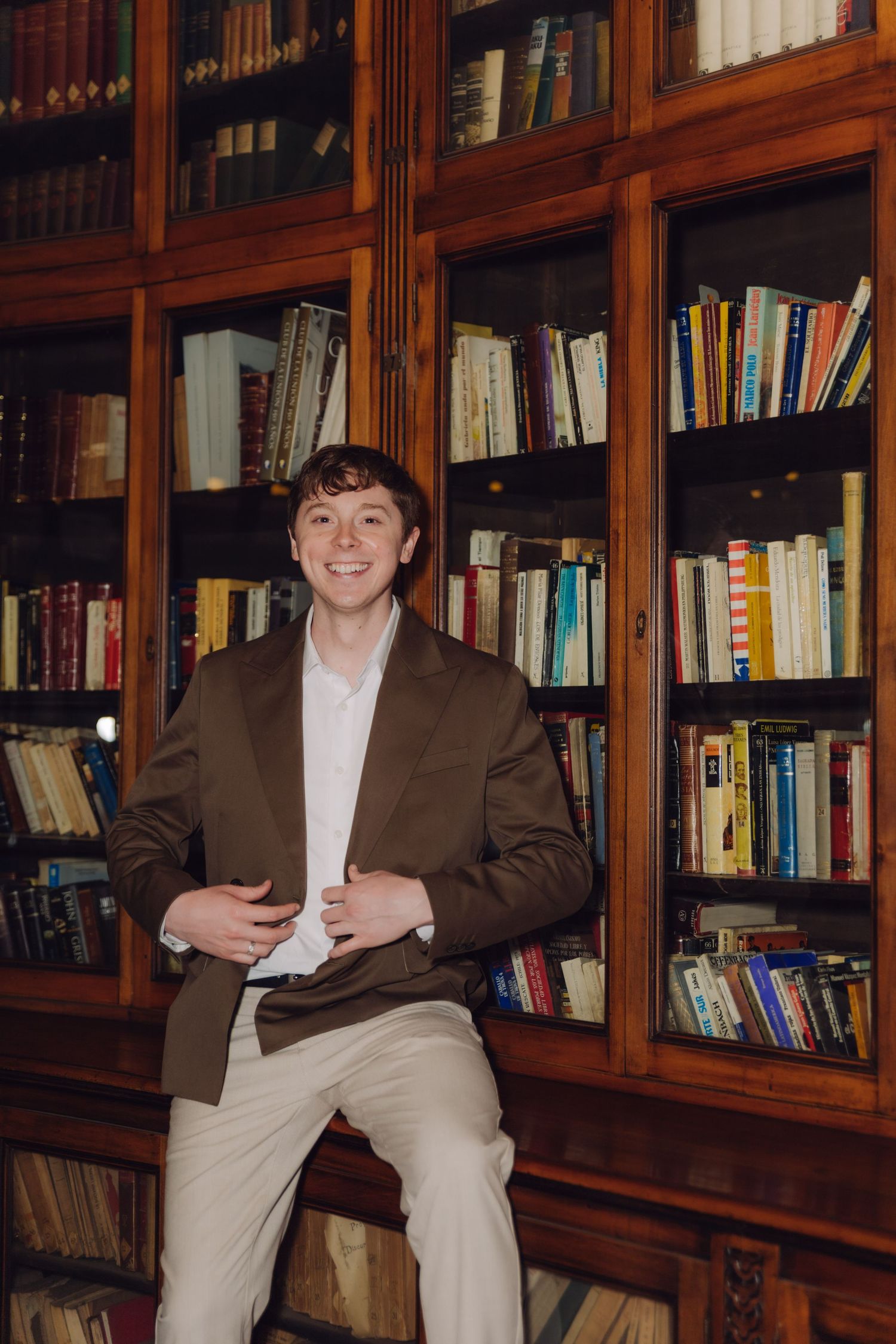 Smiling young man in brown blazer sitting against wooden library bookshelf.