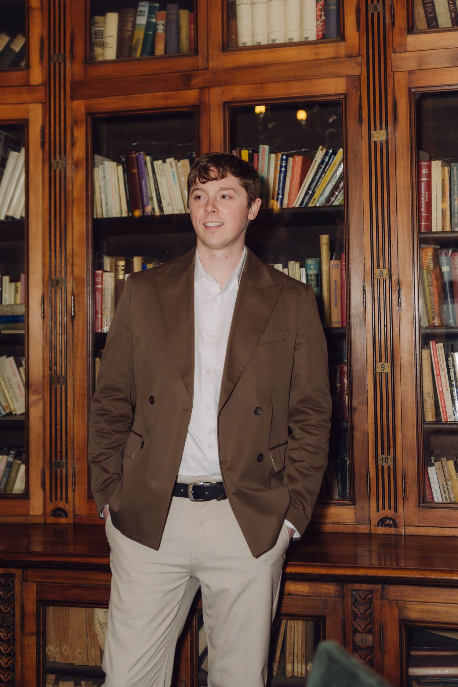 Young man in brown double-breasted blazer standing confidently in front of classic library bookcase.