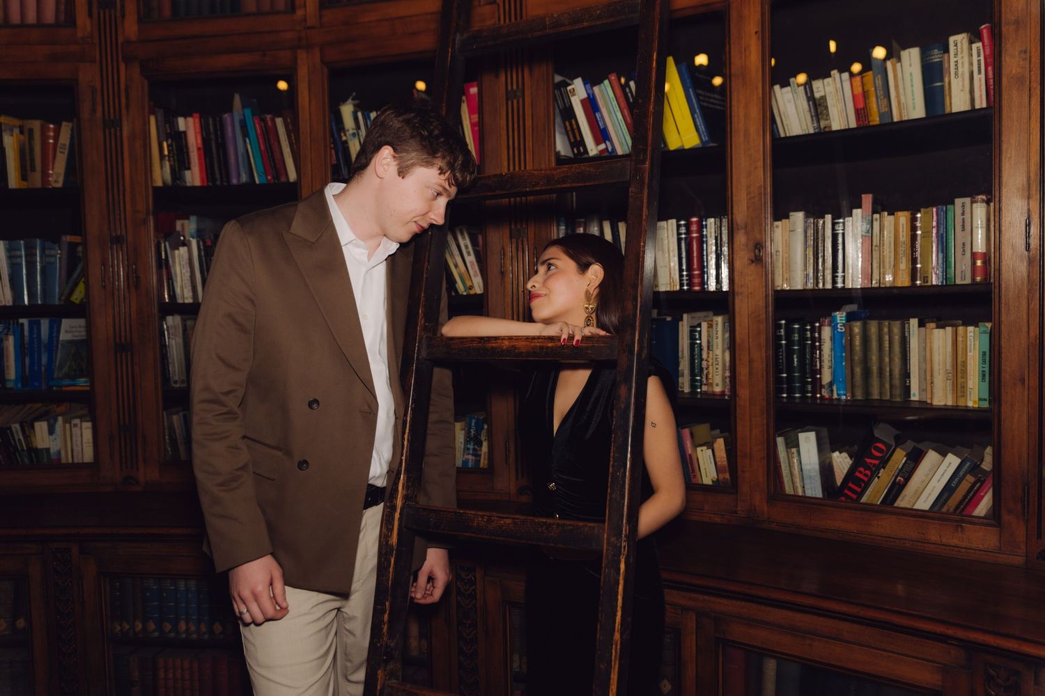 Couple gazing at each other beside wooden library ladder with bookshelves in background.