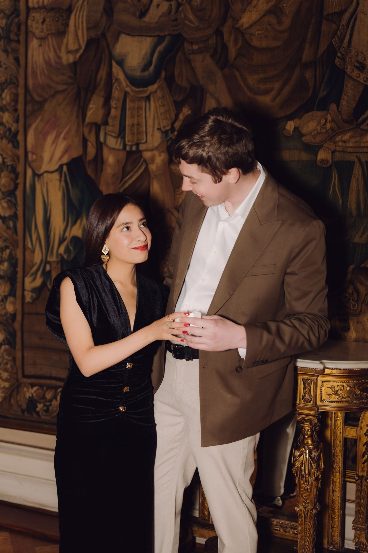 Elegant couple holding hands in ornate room with historic tapestry, woman in black velvet dress.