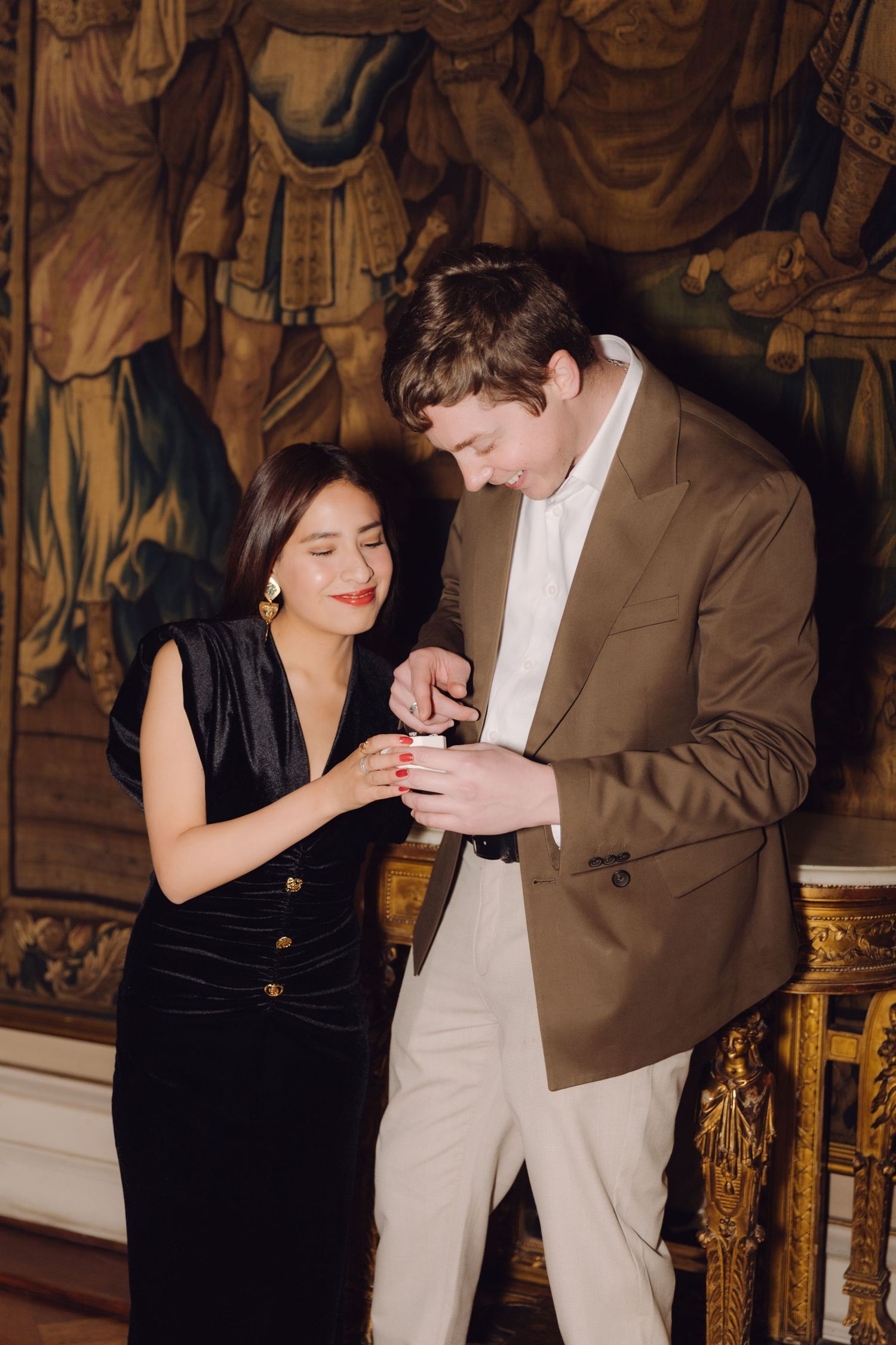 Man placing engagement ring on womans finger in elegant room with decorative tapestry backdrop.