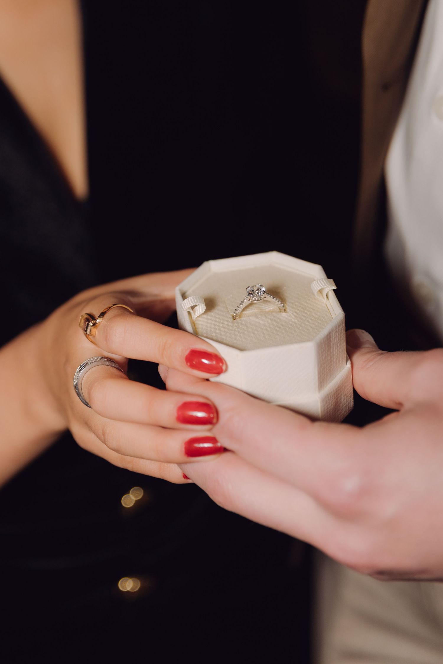Close-up of diamond engagement ring in white velvet box held by hands with red nails.
