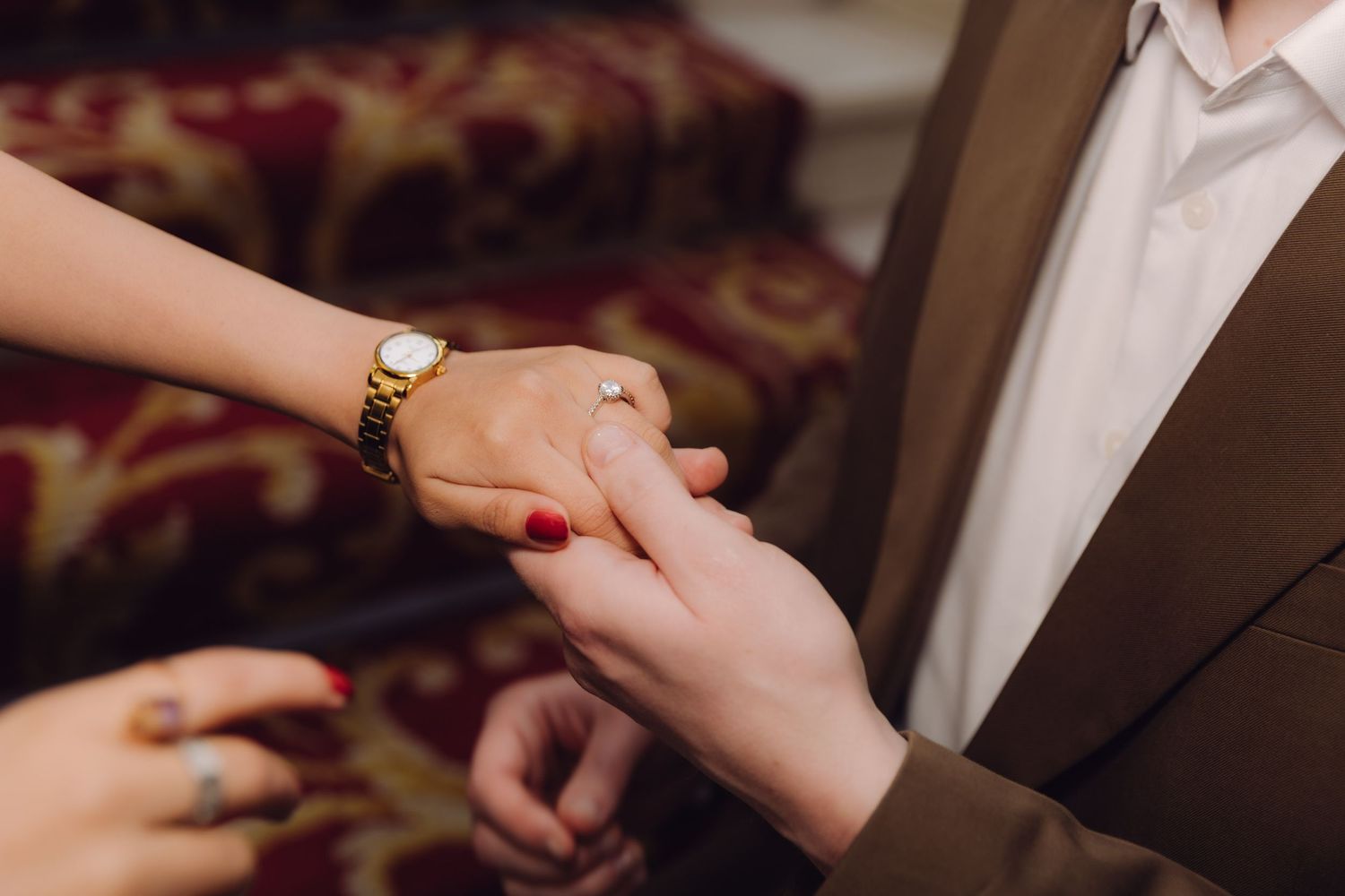 Close-up of couple holding hands with engagement ring, woman wearing gold watch and red nail polish.