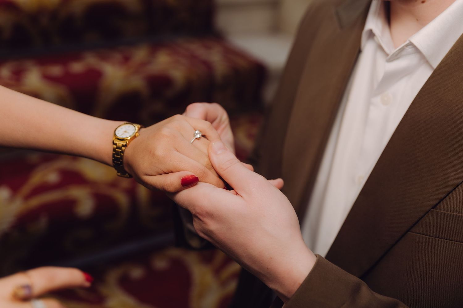 Romantic close-up of couple holding hands with diamond engagement ring and gold watch on ornate staircase.