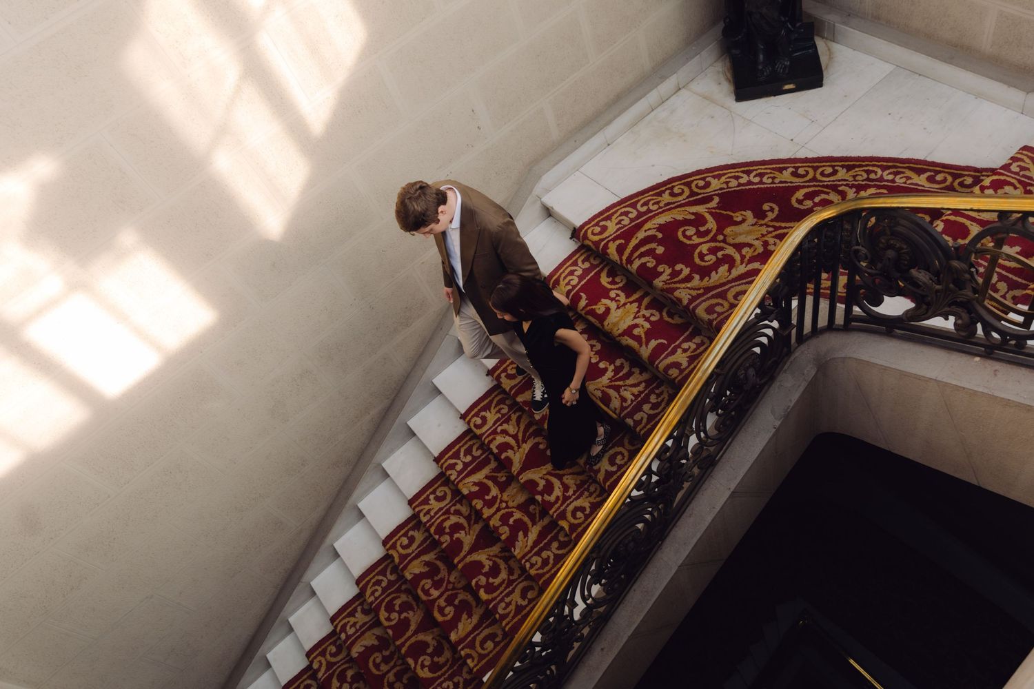 Aerial view of person in brown blazer descending grand staircase with red ornate carpet and gold railing.