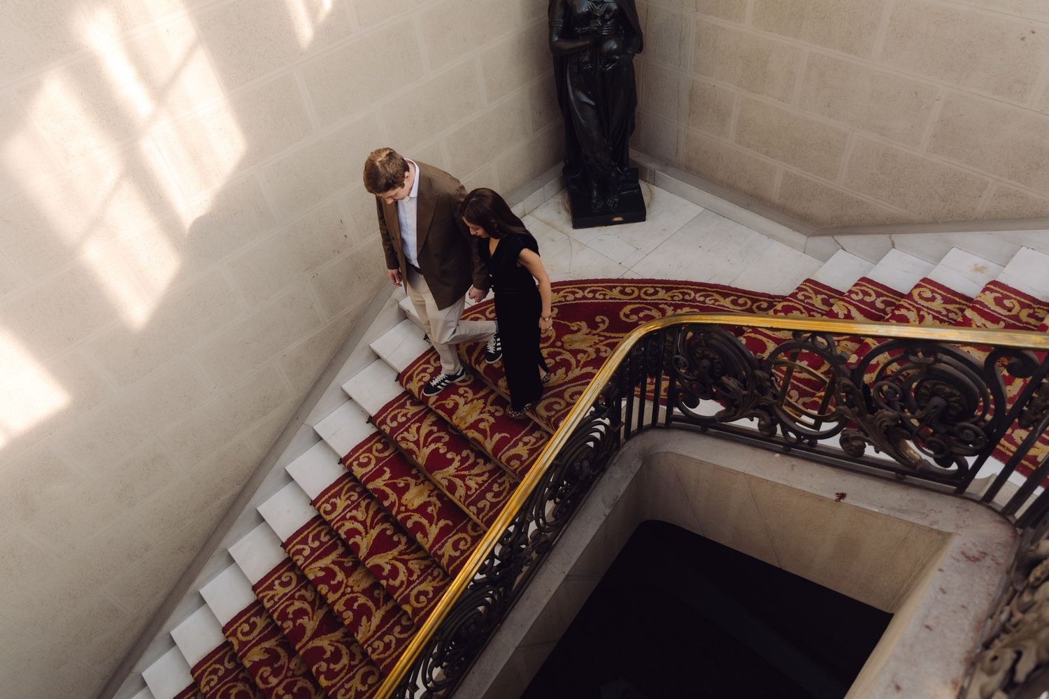 Overhead view of elegant couple on grand staircase with red patterned carpet and decorative iron railing.