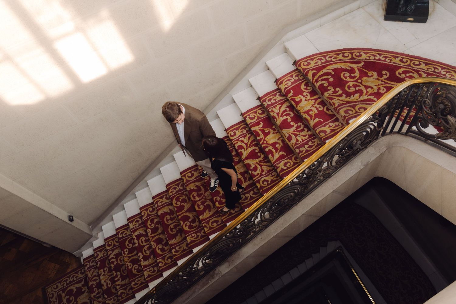 Bird's eye view of person in brown suit descending luxurious red carpeted staircase with ornate gold railing.