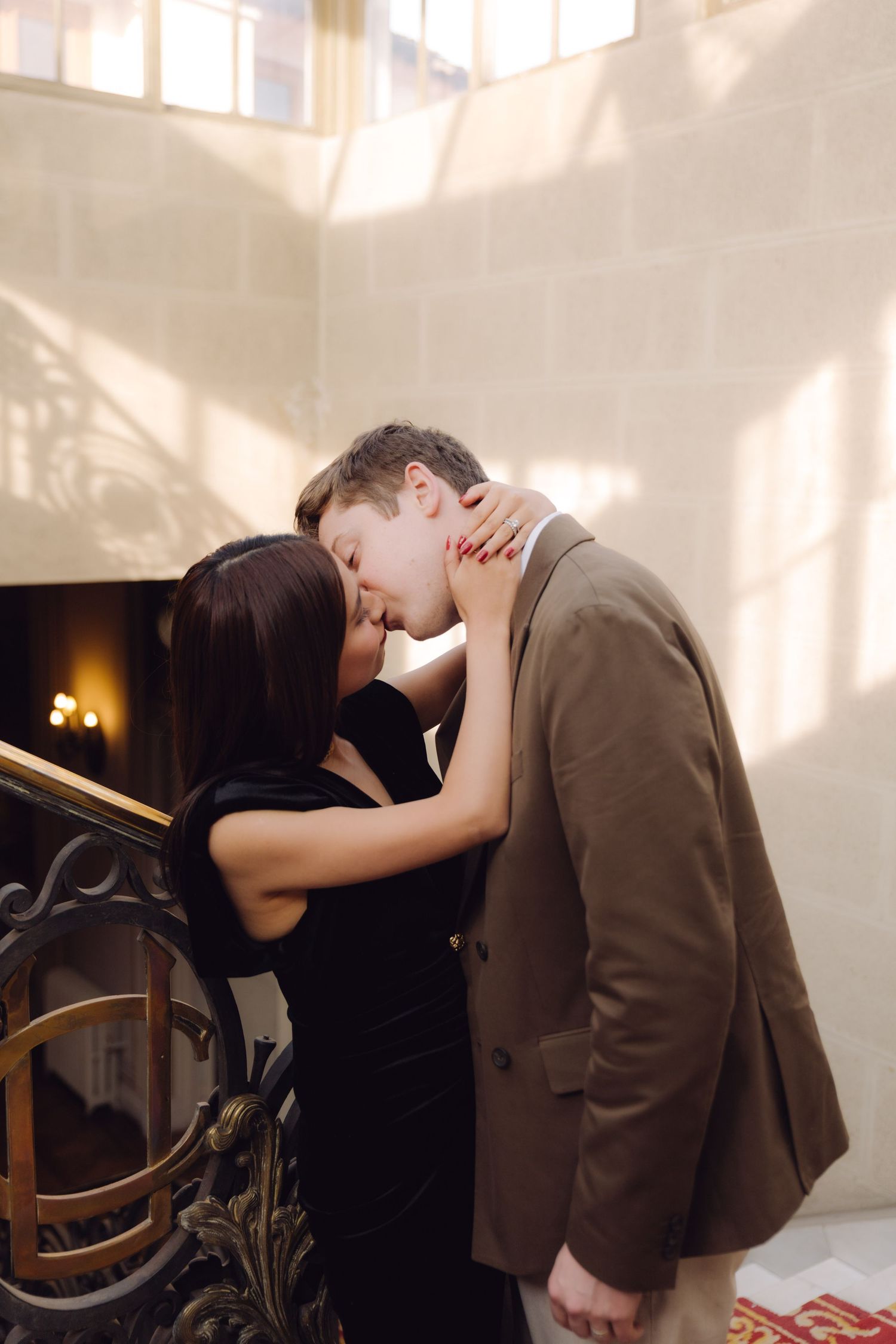 Romantic couple sharing tender kiss beside ornate gold staircase railing in elegant sunlit interior.
