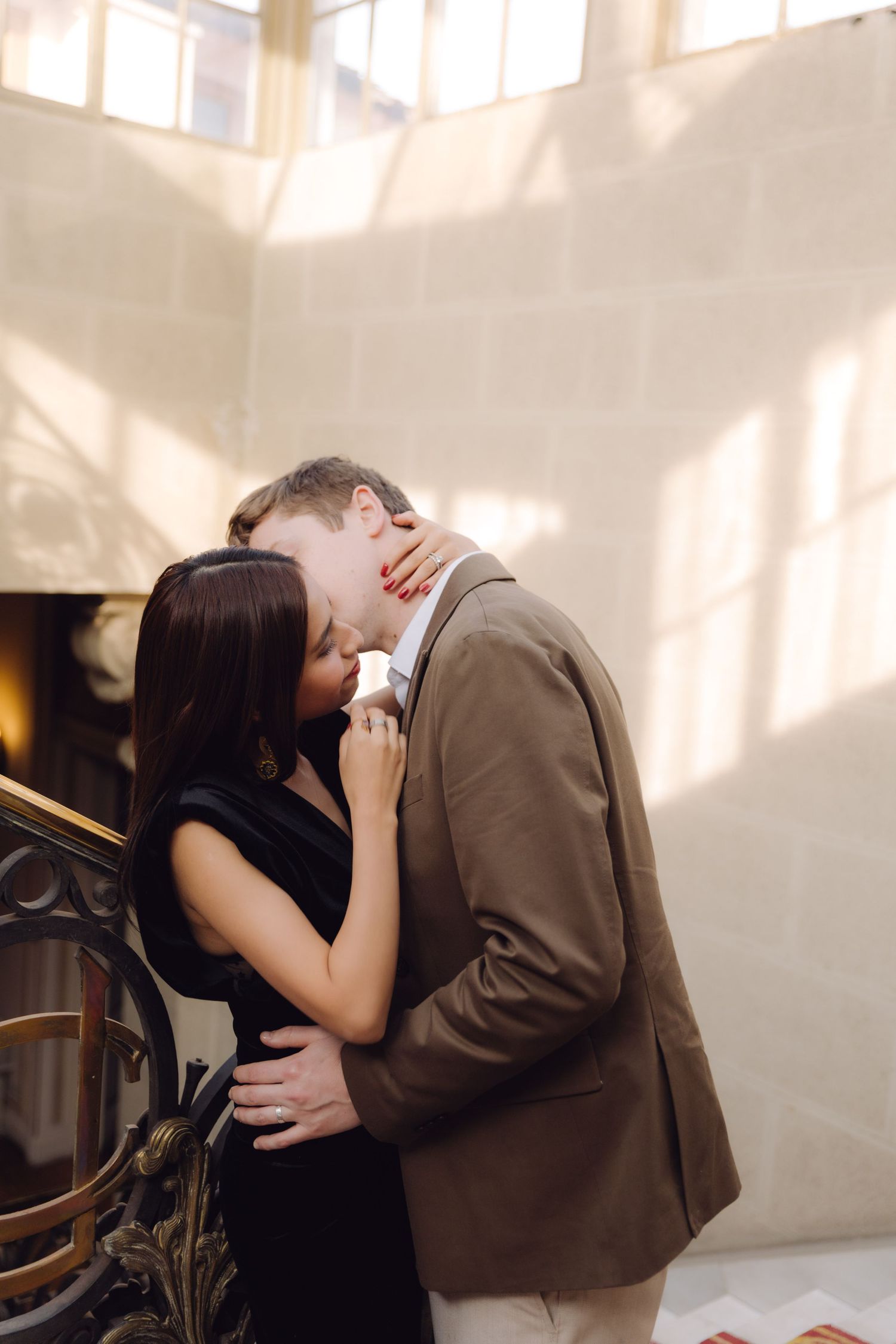 Couple embracing and kissing near decorative gold staircase banister in a bright elegant venue.