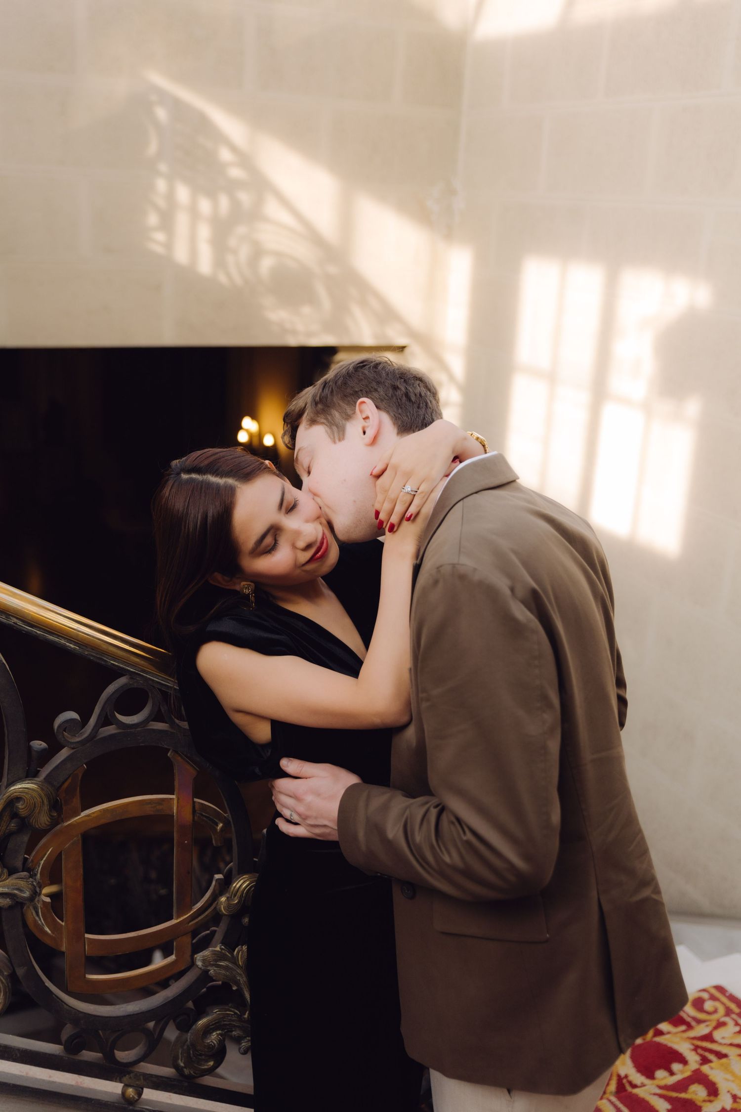 Smiling woman in black dress embracing man in brown blazer beside ornate gold staircase railing.