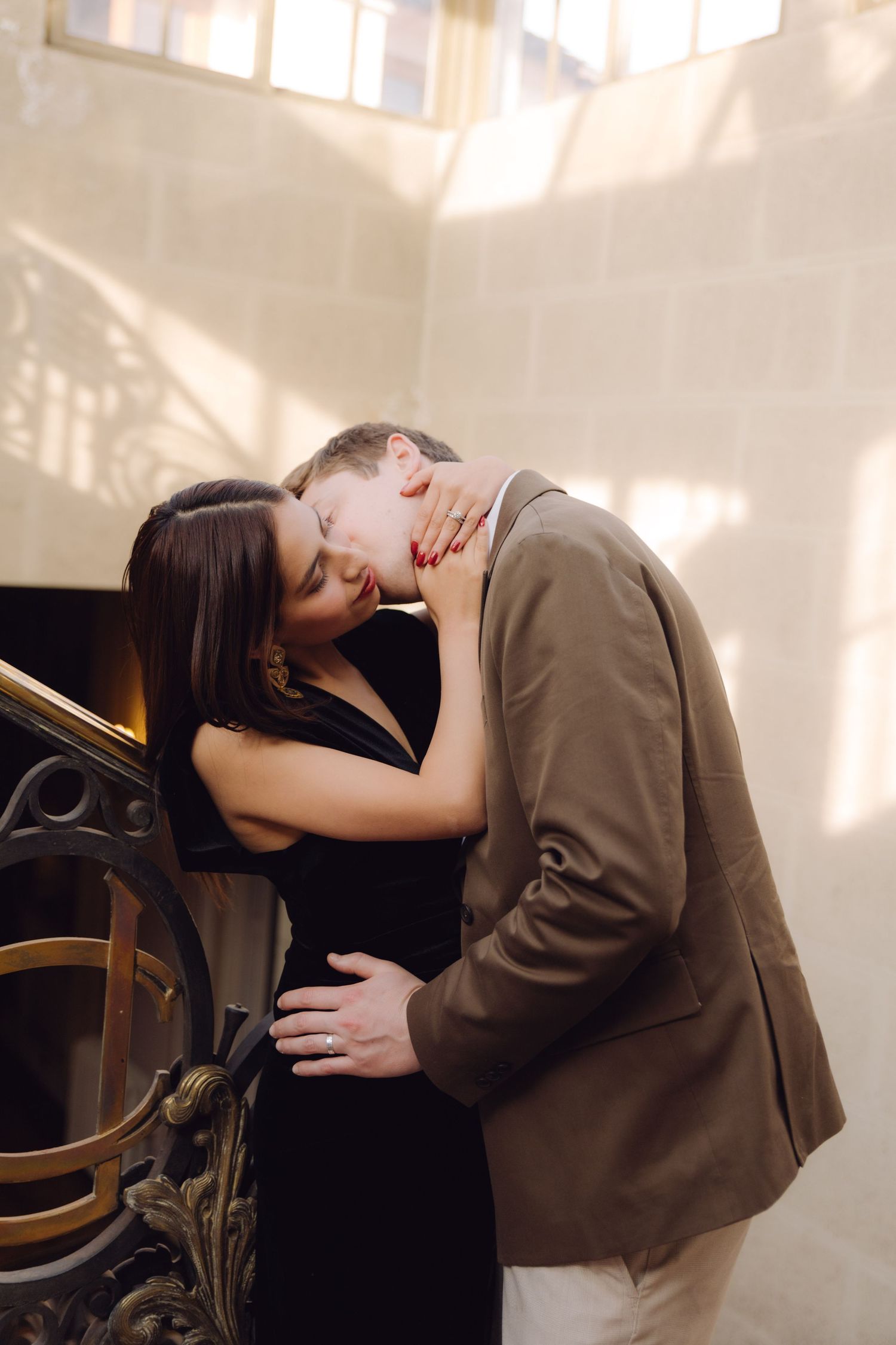 Couple sharing intimate kiss beside ornate gold staircase in sunlit elegant interior setting.
