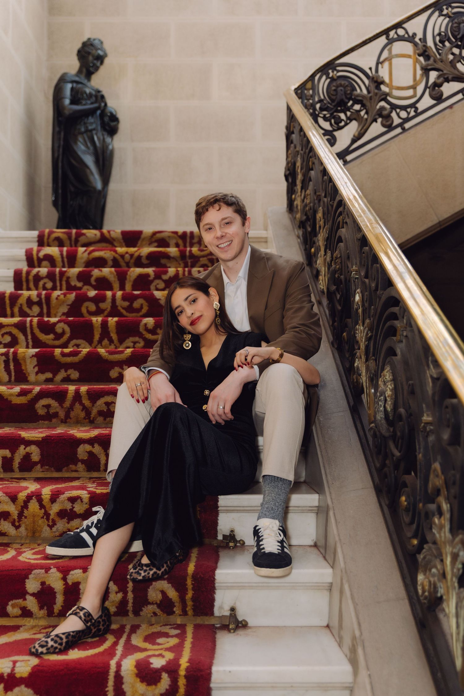 Happy couple sitting together on red ornate carpeted staircase with decorative iron railing in elegant venue.