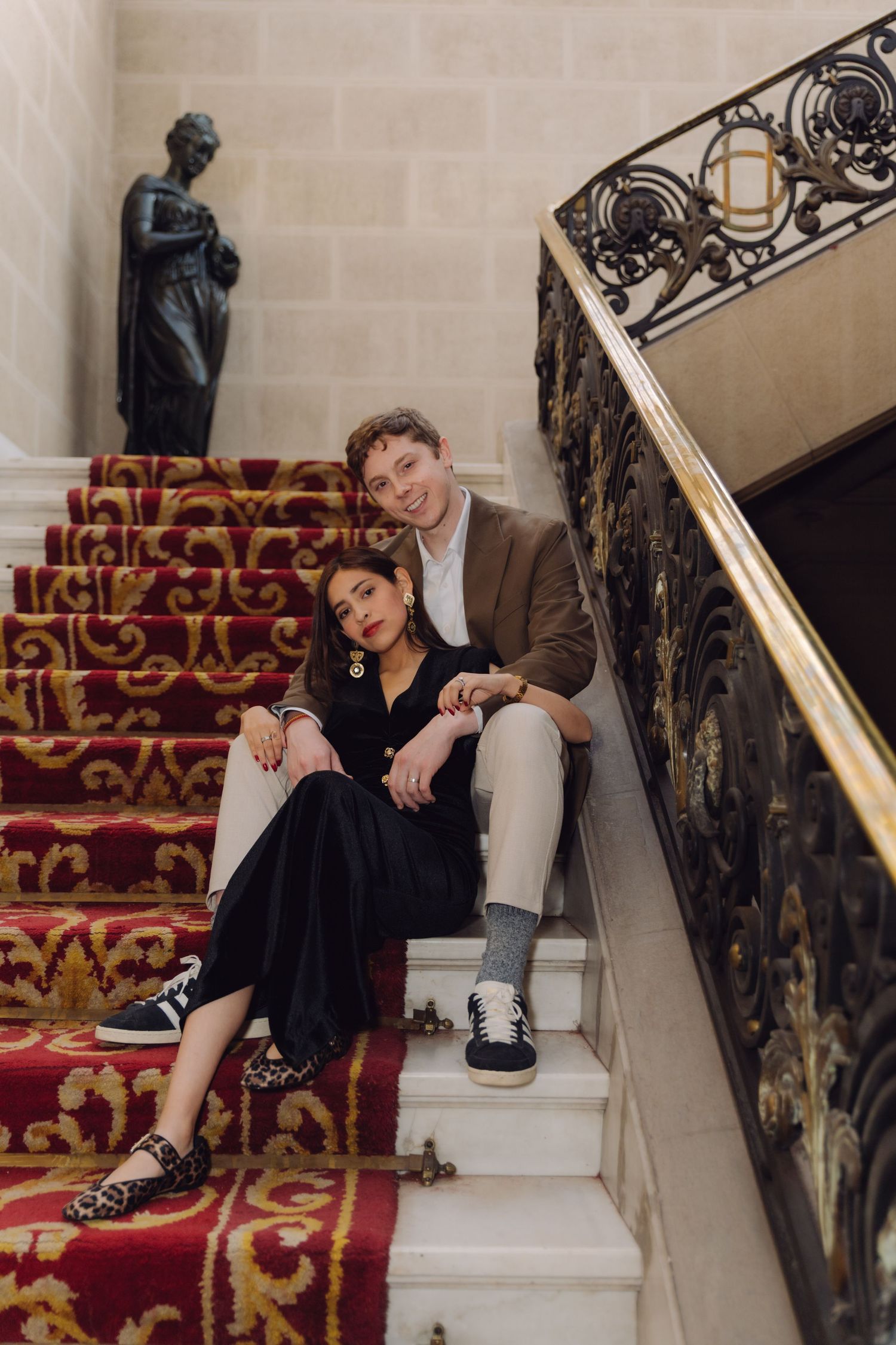 Elegant couple sitting on ornate red-carpeted stairs with wrought iron railing in a grand historic building.