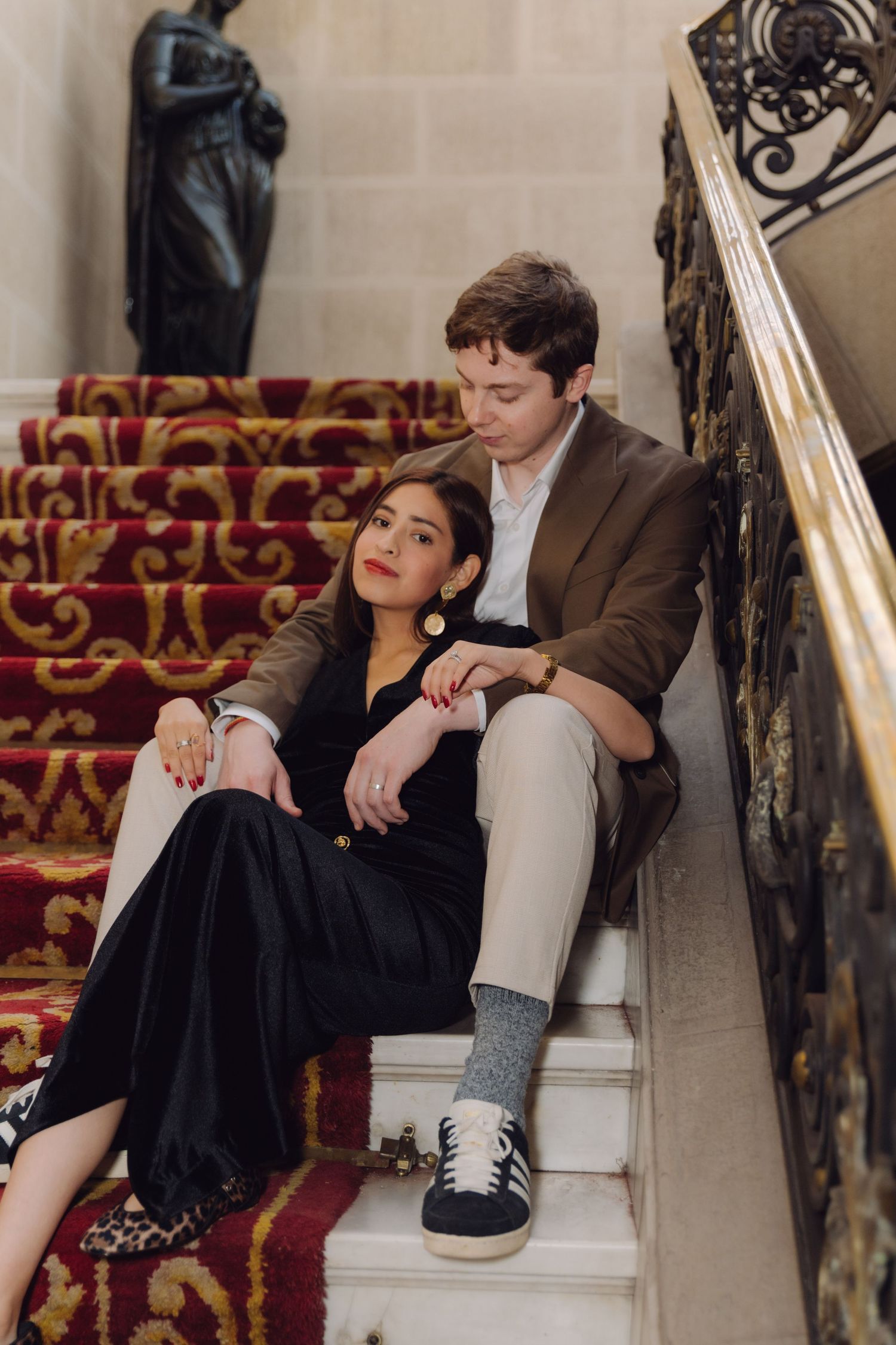 Romantic couple relaxing on red-carpeted grand staircase with decorative iron railings in luxury venue.