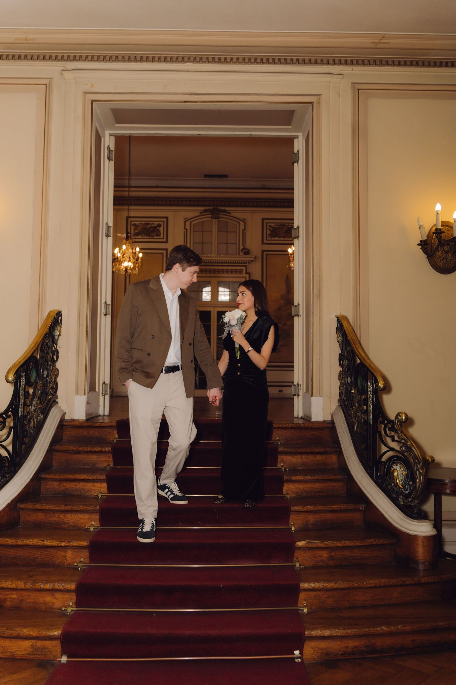 Stylish couple holding hands on red-carpeted grand staircase in elegant chandelier-lit historic mansion.