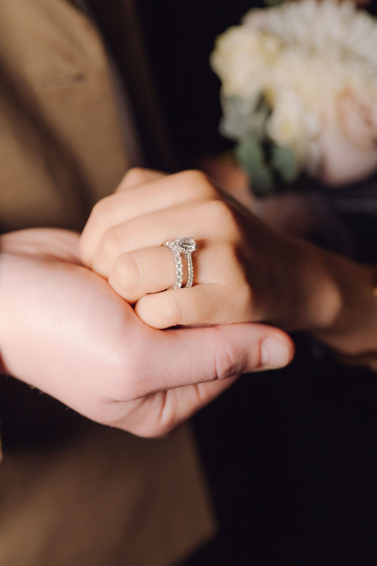 Detail shot of sparkling diamond engagement ring set on bride's finger with white floral bouquet.