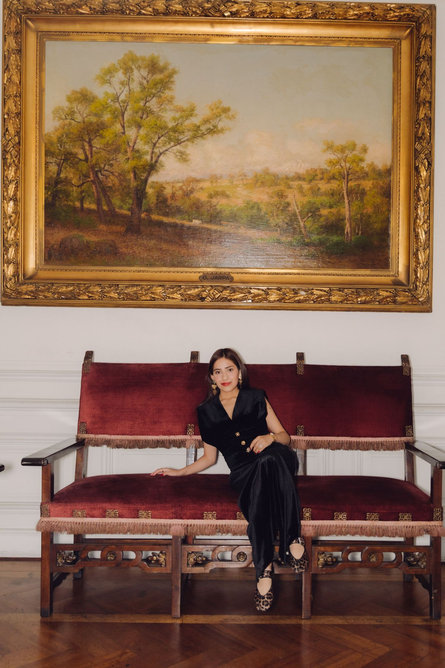 Elegant woman in black velvet dress seated on antique red velvet bench beneath gold-framed landscape painting.