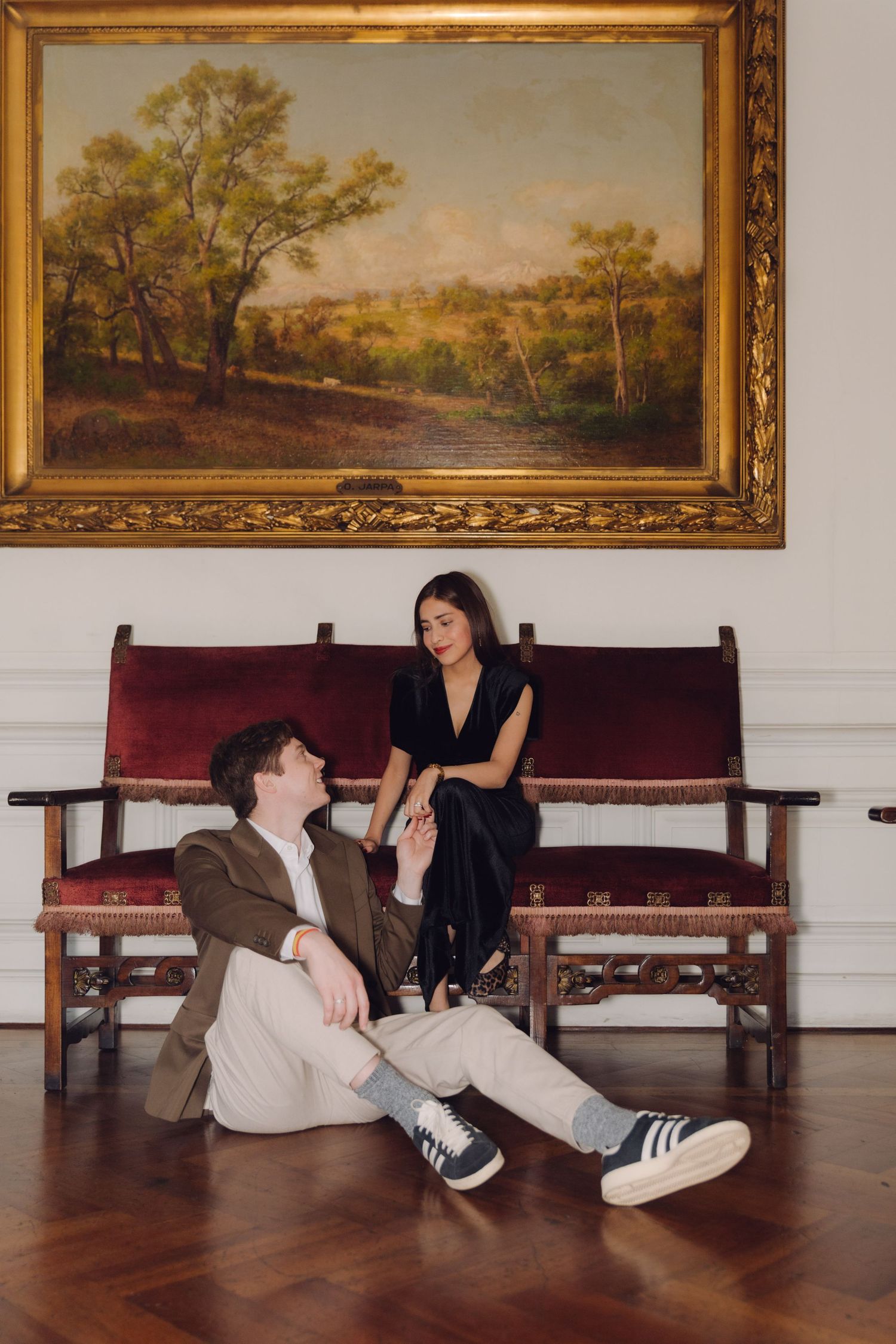 Couple posing in grand room with man sitting on floor and woman on antique red velvet bench.