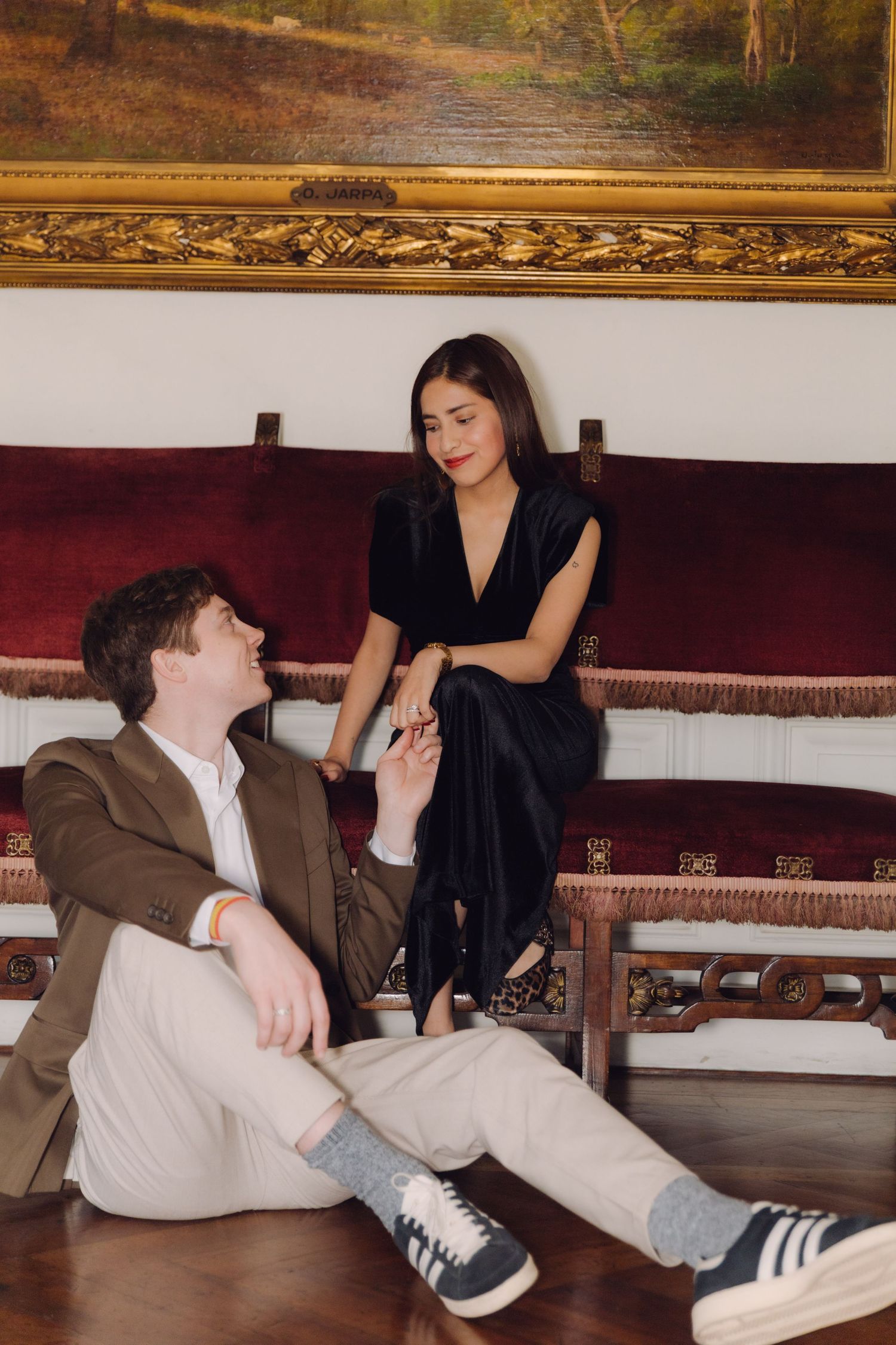 Couple holding hands as man sits on parquet floor beside woman perched on antique red velvet bench.