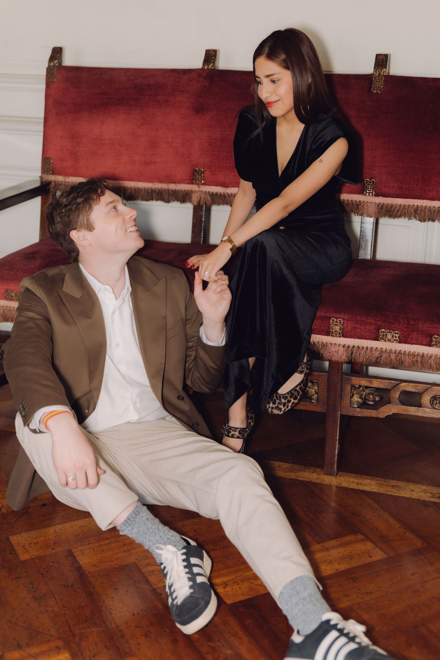 Smiling couple holding hands with man seated on wooden floor beside woman on ornate red velvet bench.