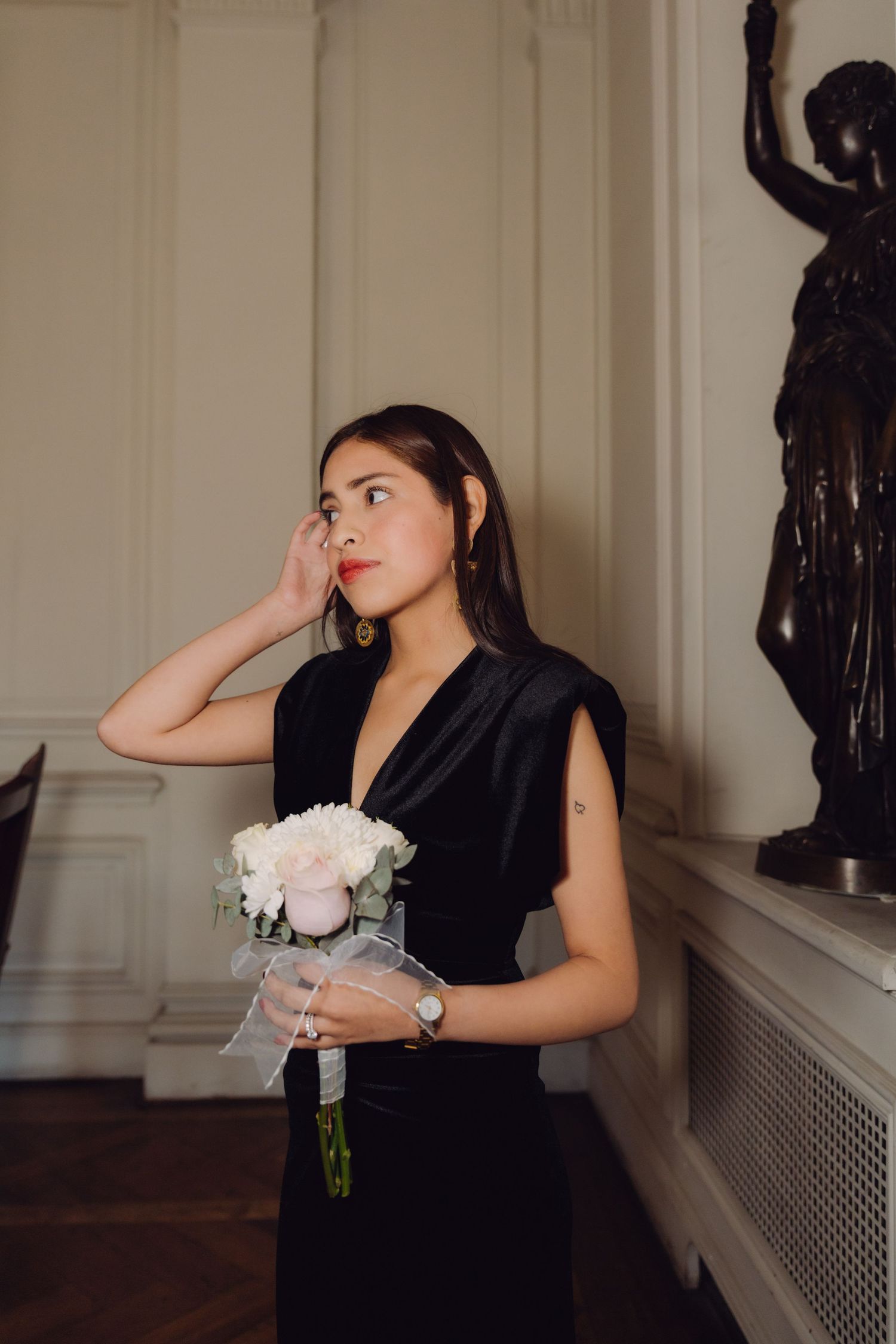 Elegant woman in black velvet dress holding white peony bouquet beside bronze statue in classic interior.