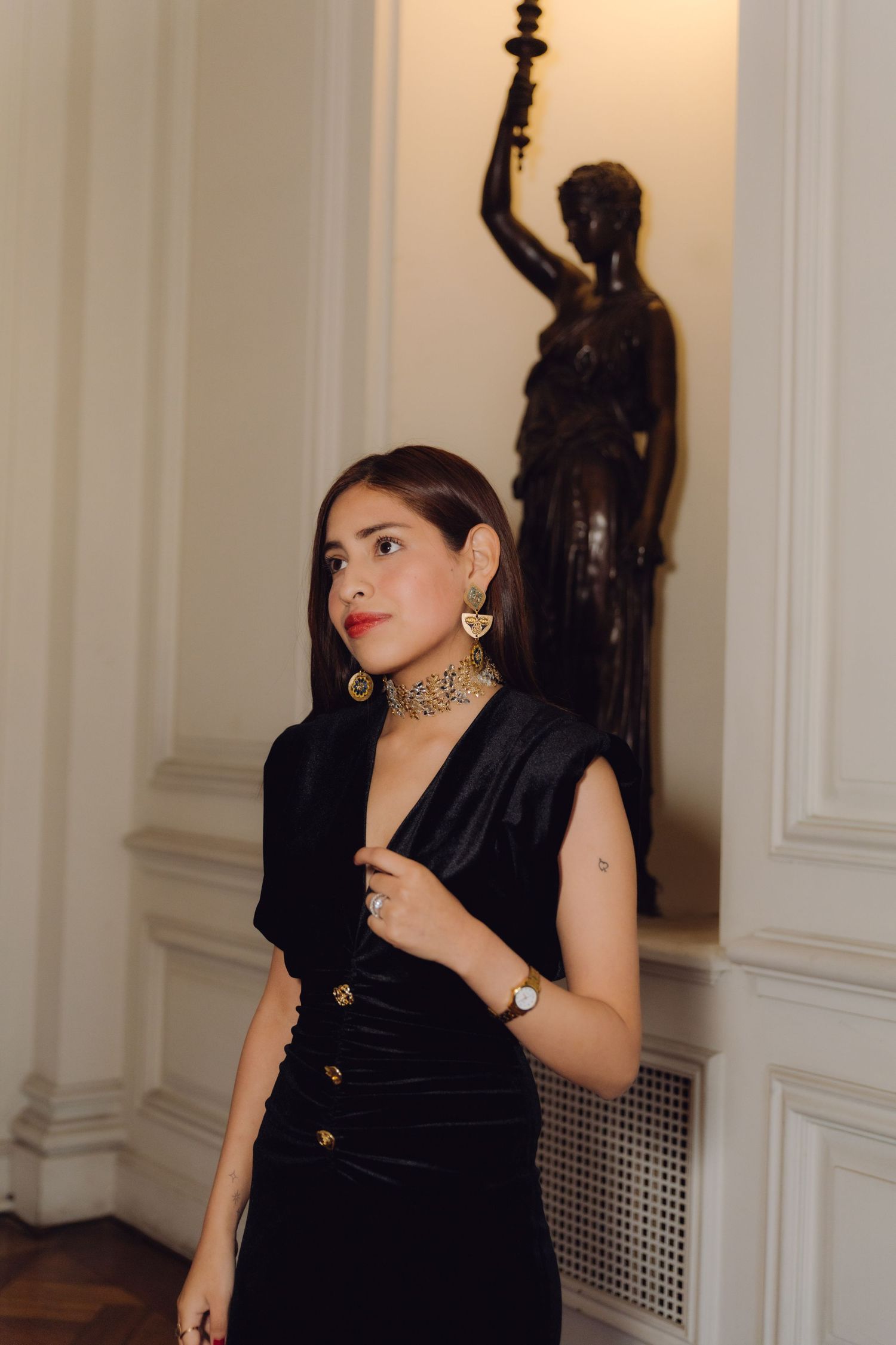 Elegant woman in black velvet dress with gold jewelry poses near classical bronze statue in ornate interior.