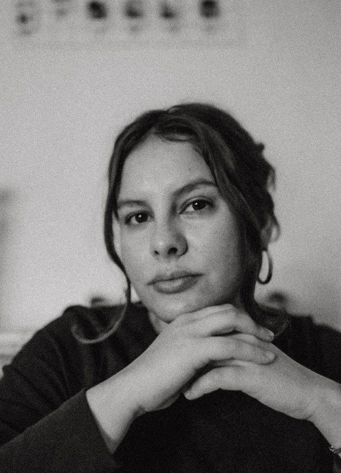 Black and white portrait of a young woman with her hands clasped under her chin, wearing hoop earrings and a dark top.