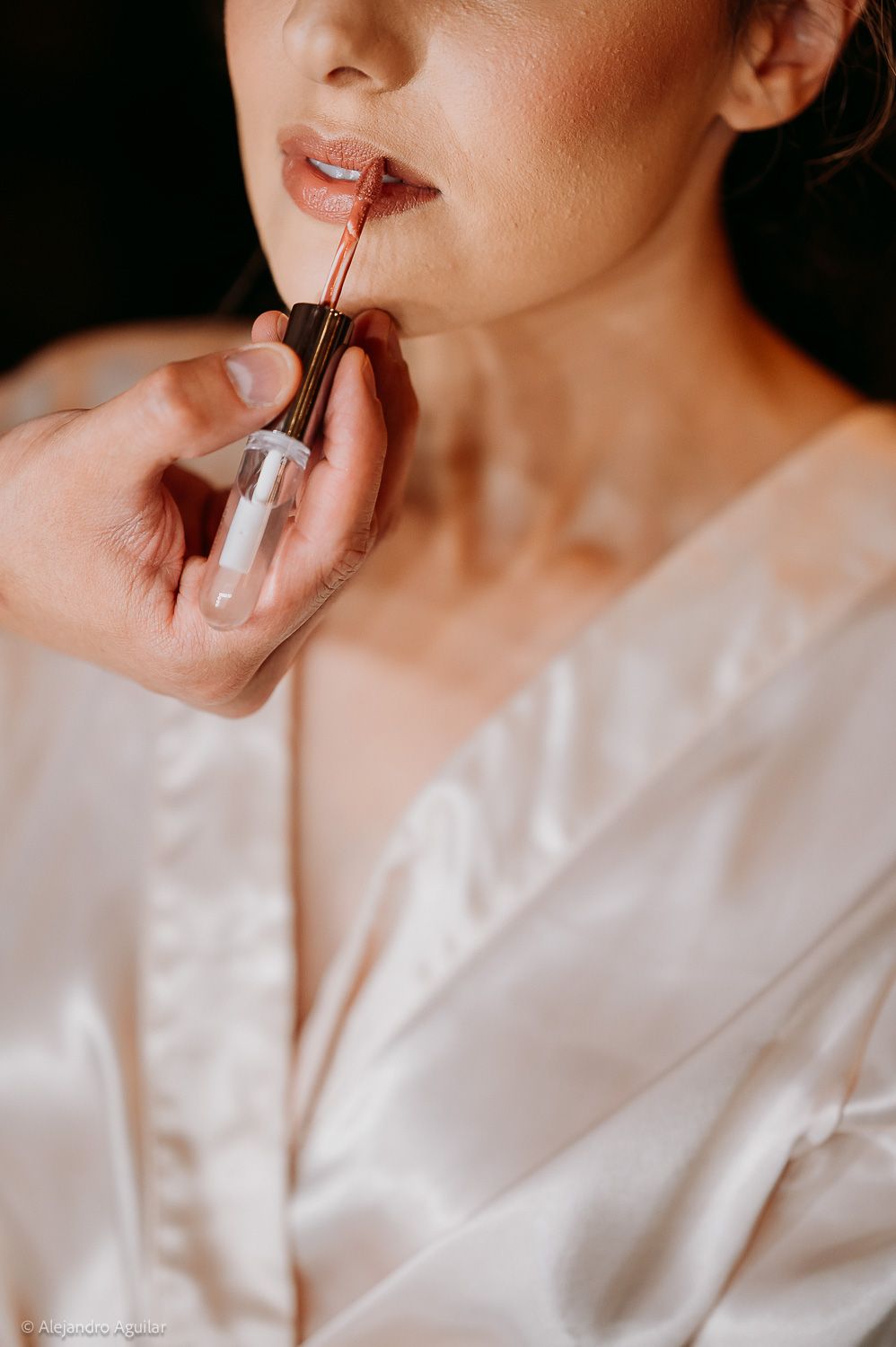 A woman in a satin robe applies nude lip gloss during bridal makeup preparation.